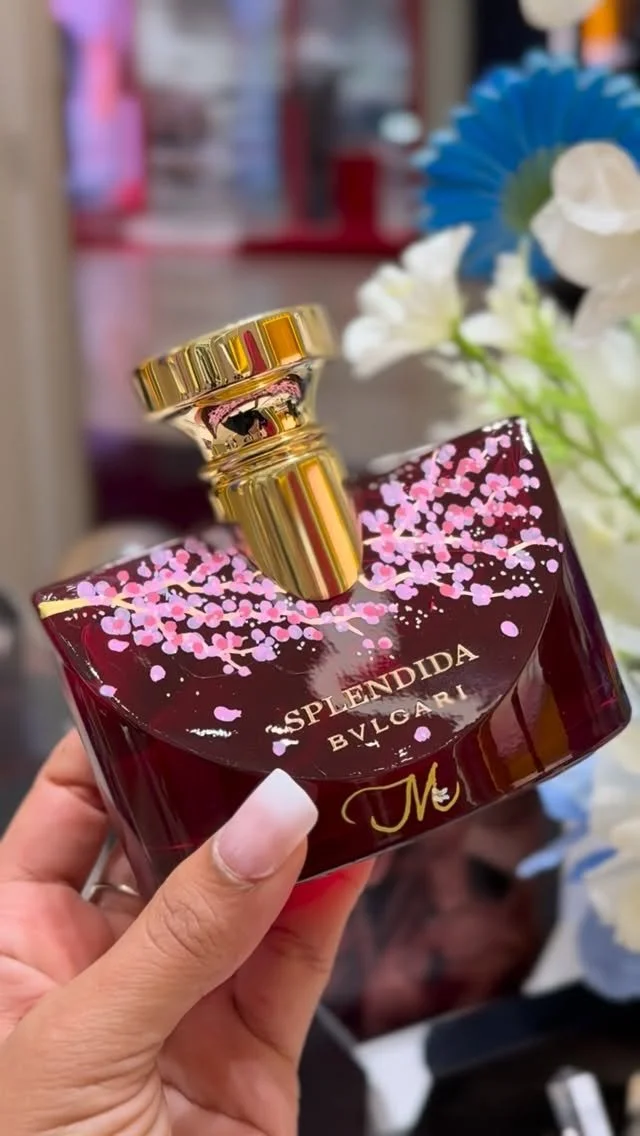 A woman holding a bottle of Bvlgari Splendida perfume with a dark red bottle and pink floral accents, with a blurred background of white and blue flowers.