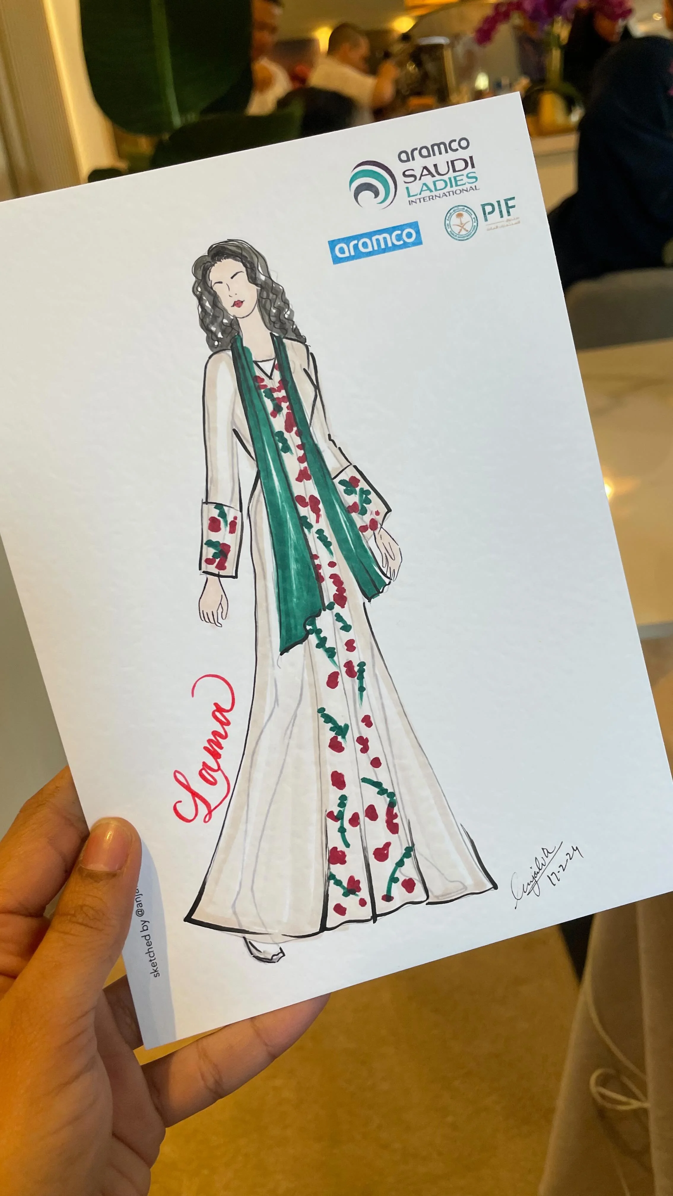 A hand holds a fashion sketch of a woman in a long dress with floral patterns and a matching scarf. The sketch features signs of sponsorship and logos from Aramco, Saudi Ladies International, and PIF, indicating a connection to a Saudi women’s golf e