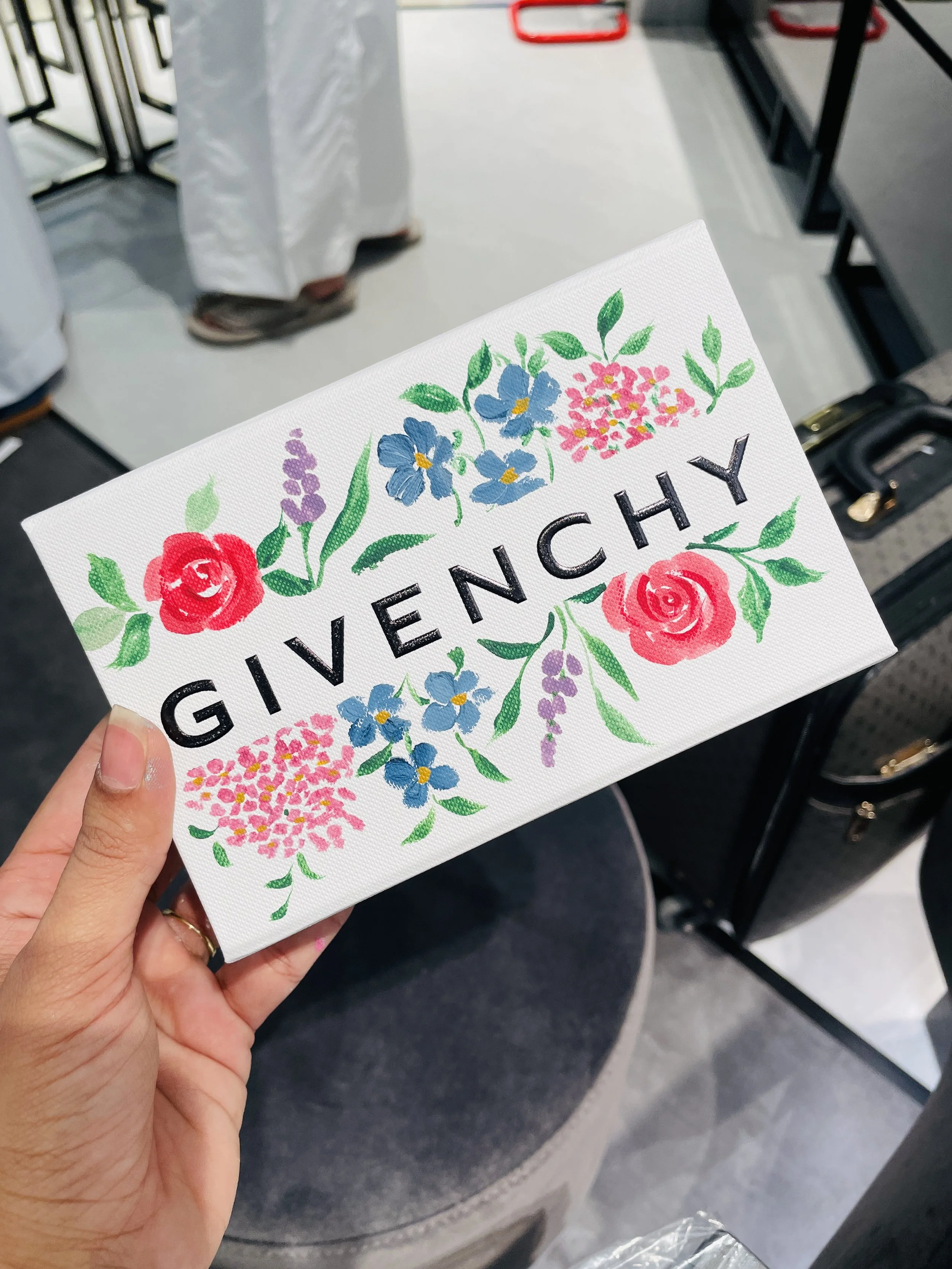 A person's hand holding a Givenchy paper bag with floral design, showing blue, pink, and purple flowers and green leaves.