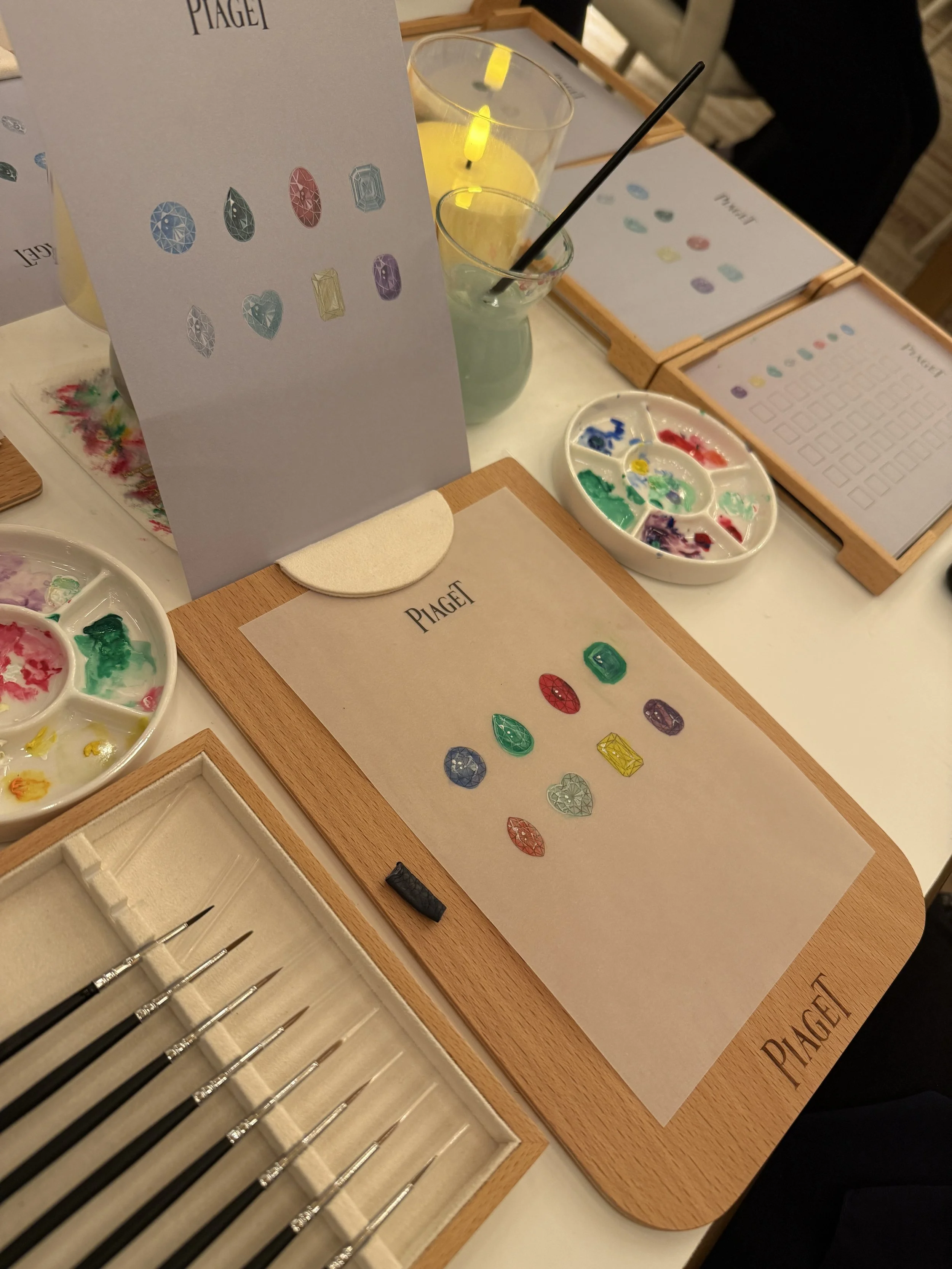 A display of colorful gemstone stickers on paper, with watercolor paint palettes, paintbrushes, and a lit yellow candle on a table labeled 'PIAGET'.