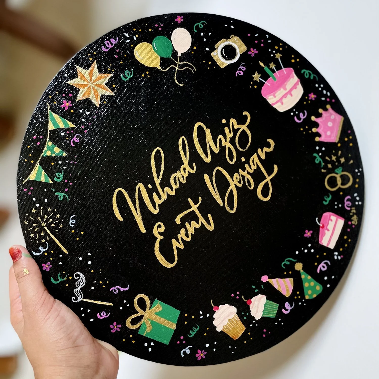 Black circular cake with gold writing saying "Michaela Ennt & Benjamin." The cake is decorated with colorful illustrations of balloons, a camera, a birthday cake, a cupcake, presents, party hats, confetti, streamers, and fireworks, indicating a celeb