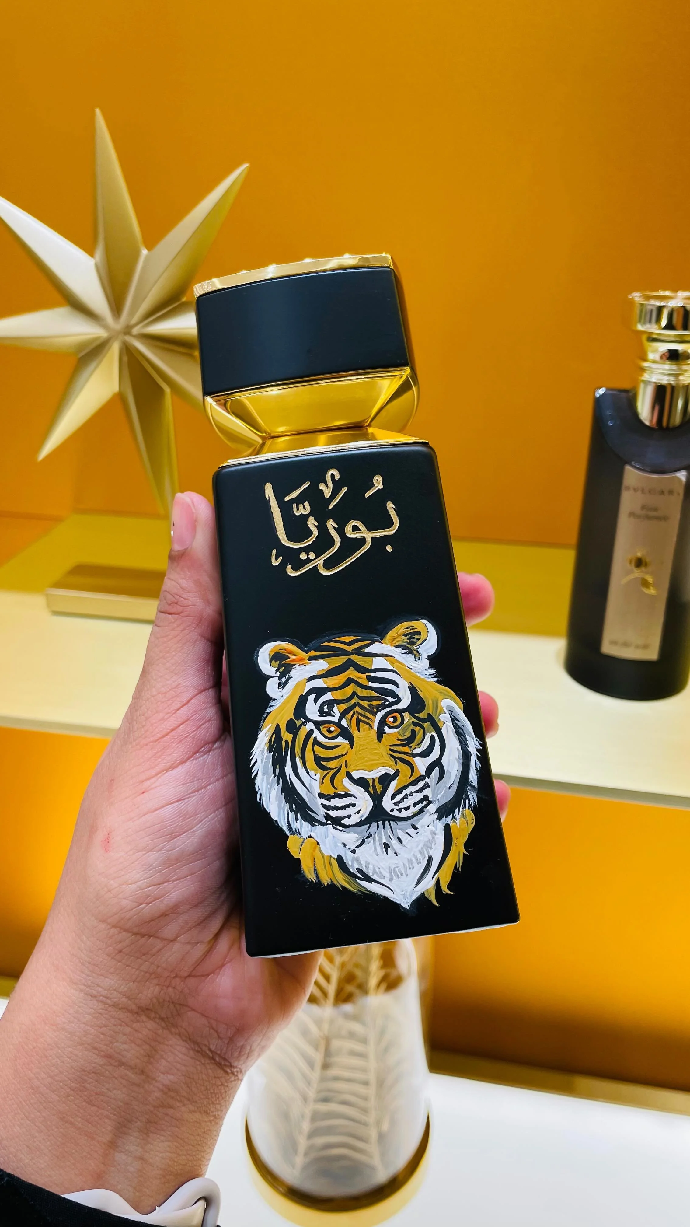 A hand holding a black and gold perfume bottle with a tiger illustration and Arabic script, in front of a yellow background with a decorative star-shaped ornament.