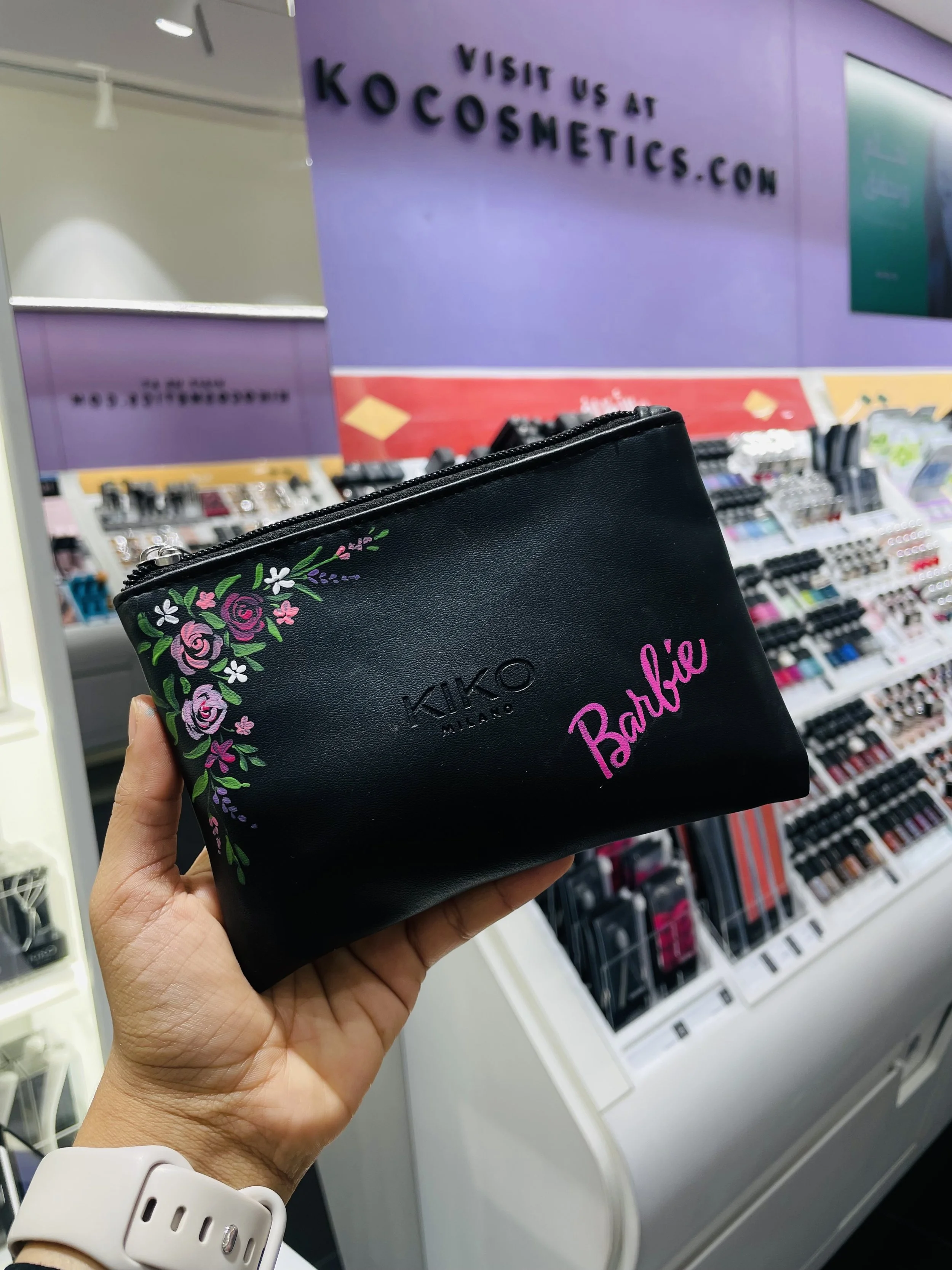 A person is holding a black KIKO Milano cosmetic pouch with floral embroidery and pink text that says 'Barbie'. They are in a store aisle with makeup products and a purple sign that reads 'Visit us at kocosmetics.com'.
