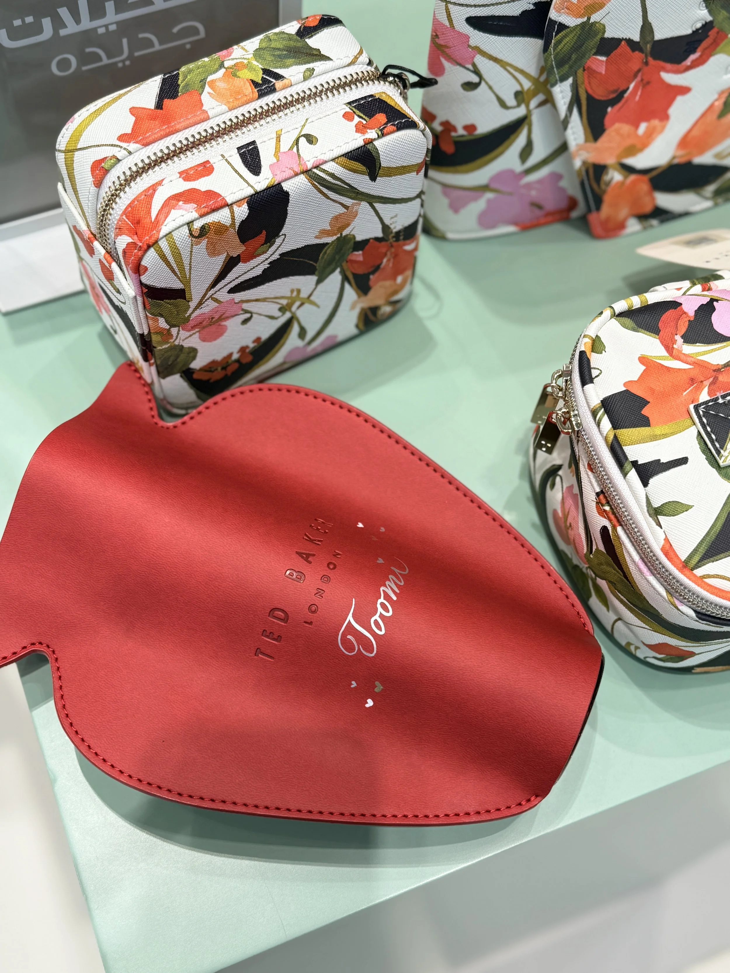 A floral-patterned small bag with a zipper, placed on a mint green round table, alongside part of a red bag with the words 'TED BAKER LONDON' and 'TOMI' written on it. In the background, there are more floral-patterned bags of similar style.