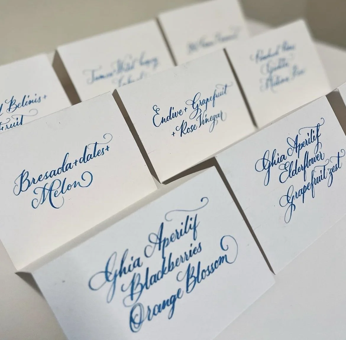 Close-up of white paper tags with elegant blue cursive writing listing names of ingredients and flavors, including rose vinegar, elderflower, blackberries, and orange blossom.