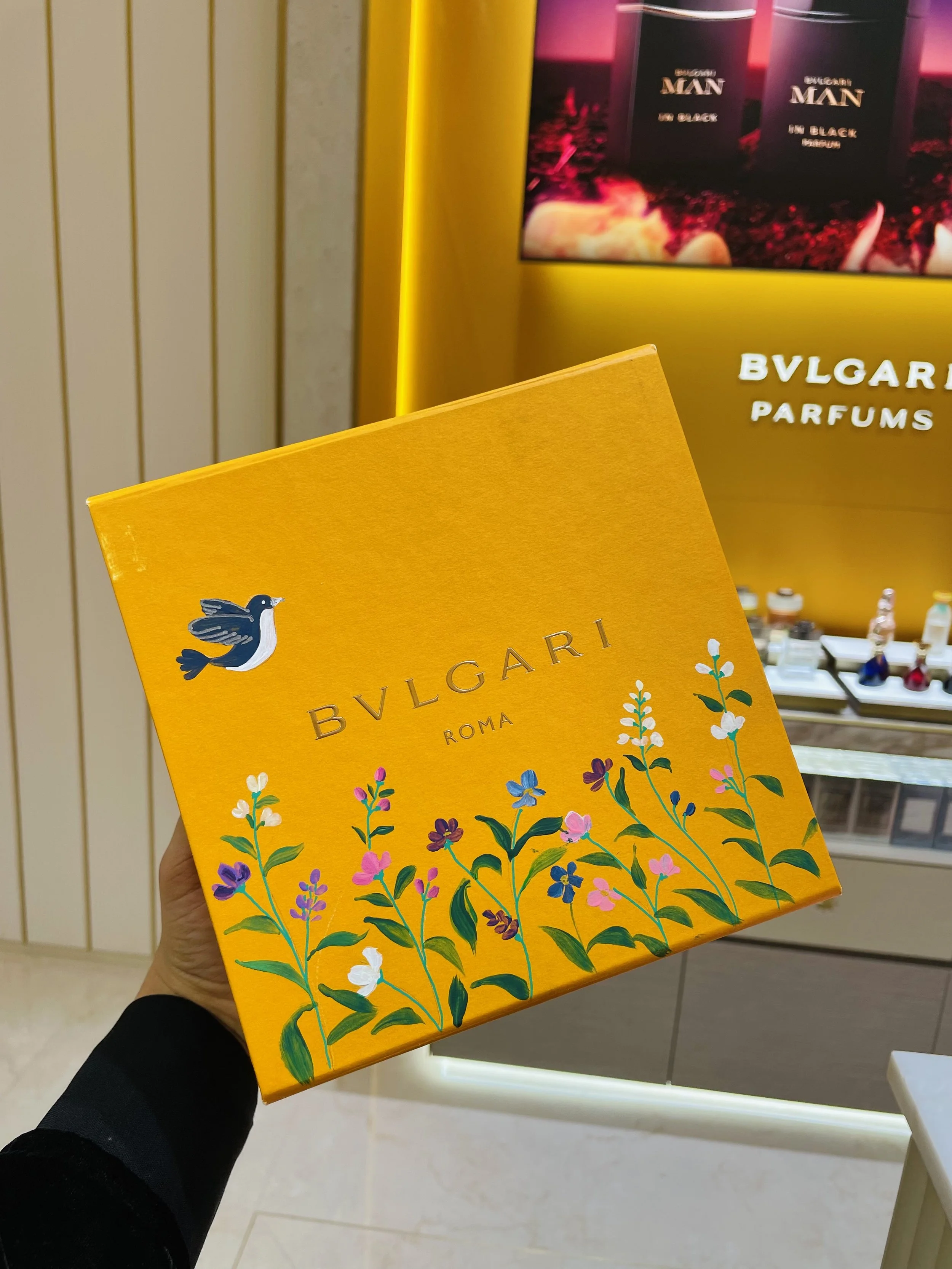 Hand holding a yellow box with floral illustrations and the brand name BVLGARI on it, with a display of BVLGARI perfumes in the background.