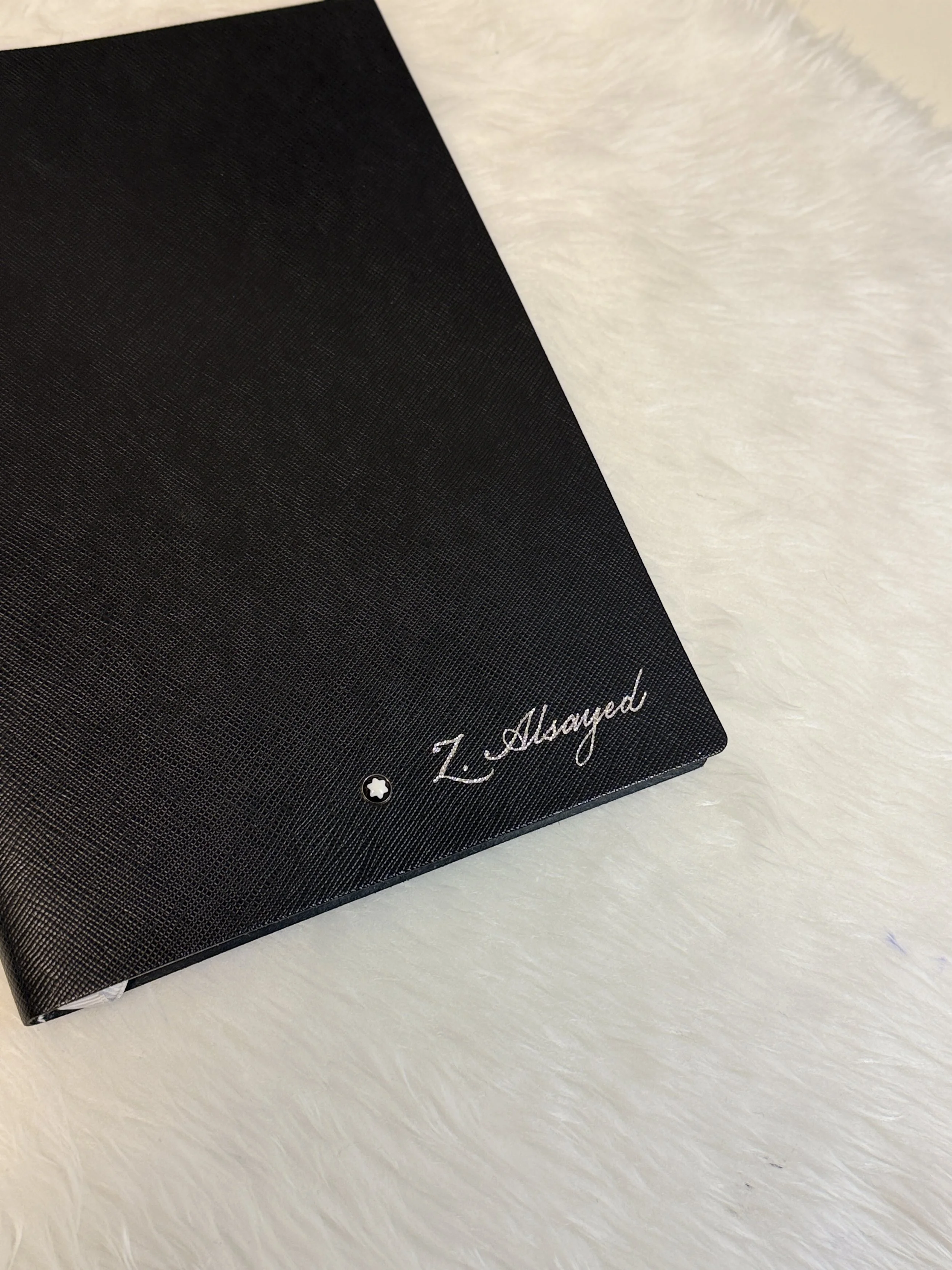 A black notebook or journal with a textured cover and the words 'I. Meayed' written in cursive in the bottom right corner, lying on a white textured surface.