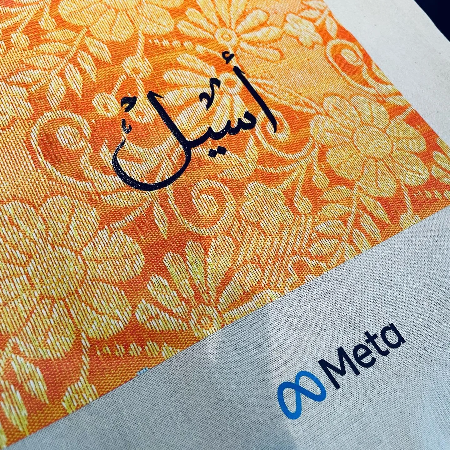 Close-up of an orange banknote with floral patterns and a handwritten signature in black ink, partly covering a document with the blue Meta logo.