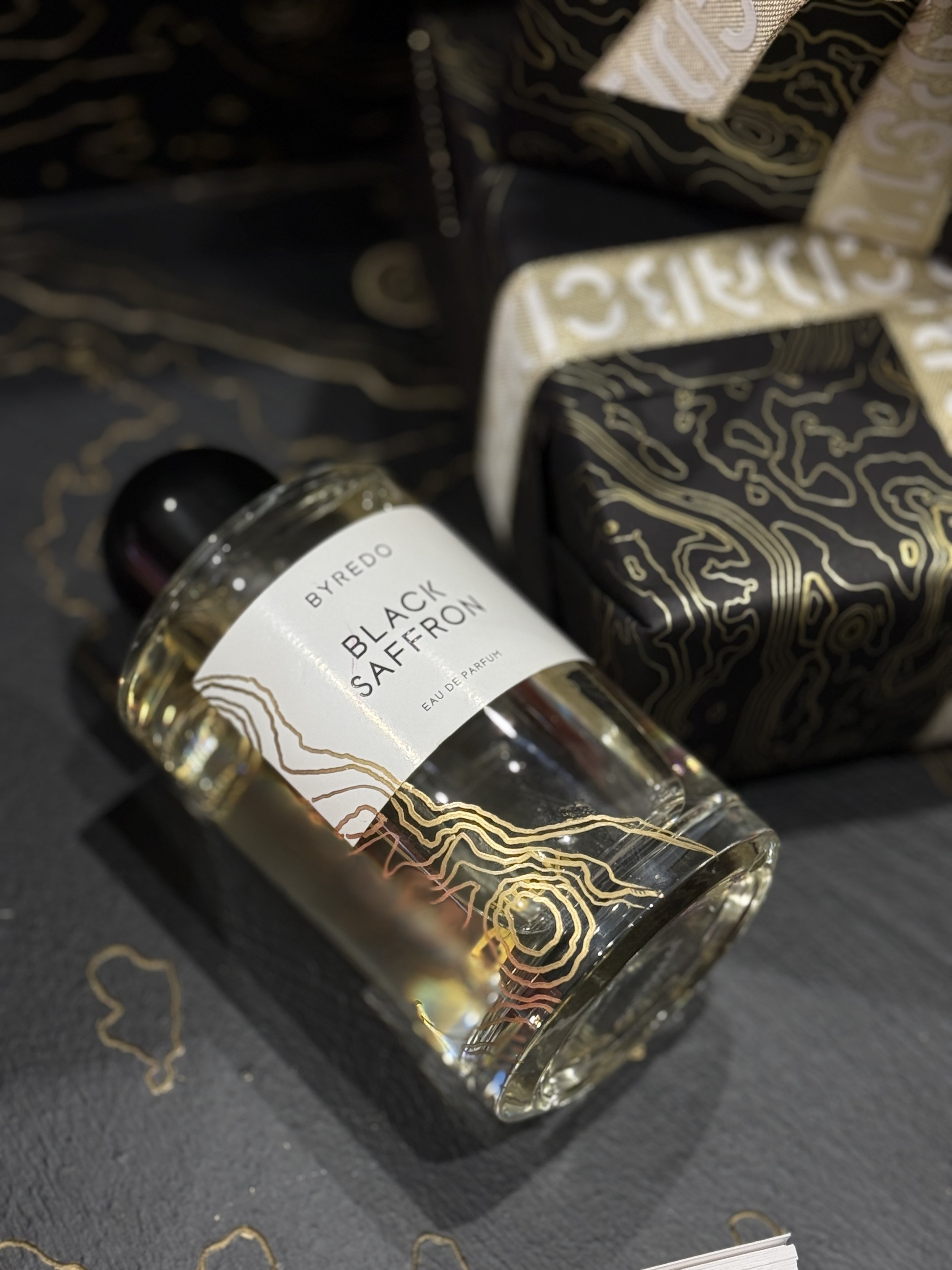 Close-up of a perfume bottle labeled 'Black Saffron' by BYREDO, with a patterned black and gold box behind it, placed on a dark surface.