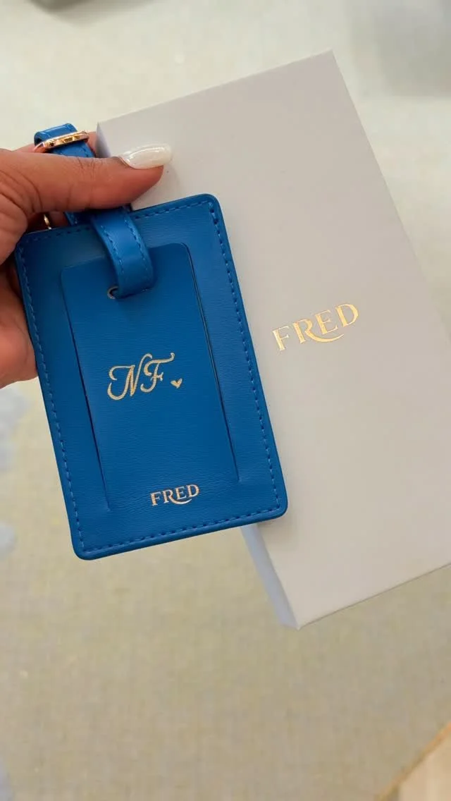 A blue FRED luggage Tag with initials 'NF' and a small heart on a person's hand, next to a white box with the FRED logo.