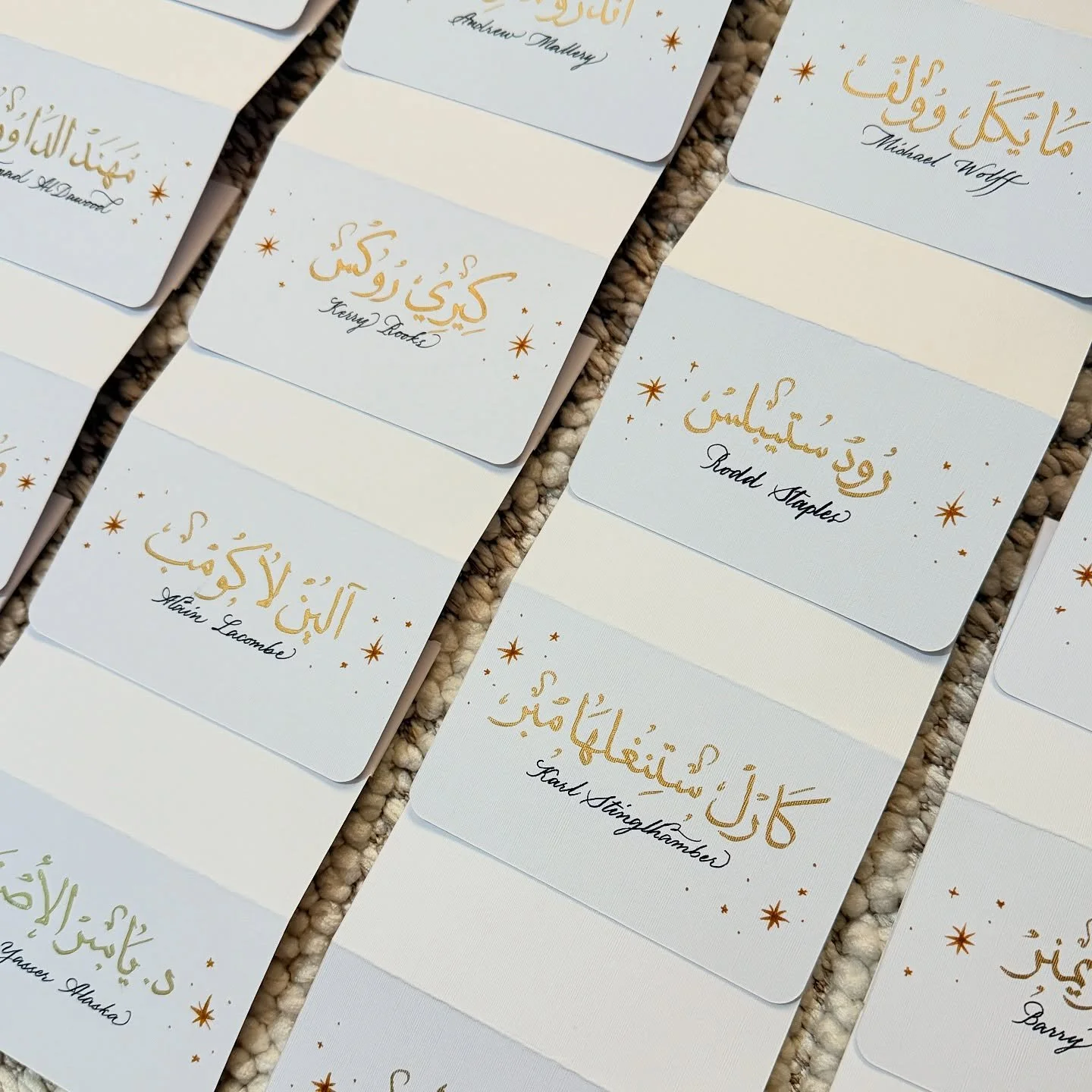 Several wedding place cards with Arabic calligraphy in gold and black, decorated with stars, on a textured beige surface.