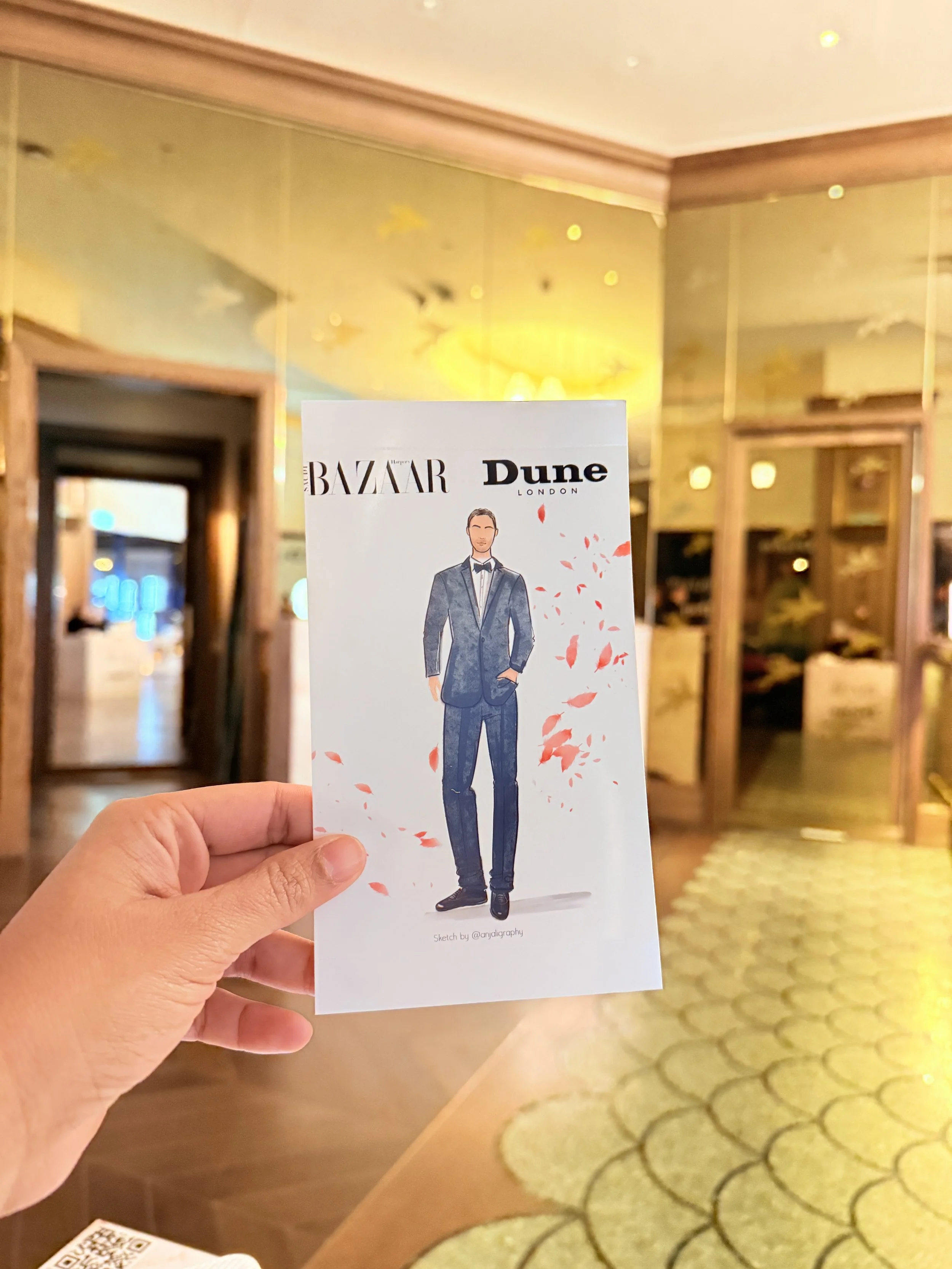 A hand holding a illustrated card of a man in a suit with the logos of Harper's Bazaar and Dune London in the background of a hotel lobby with warm lighting and wood framed doors.