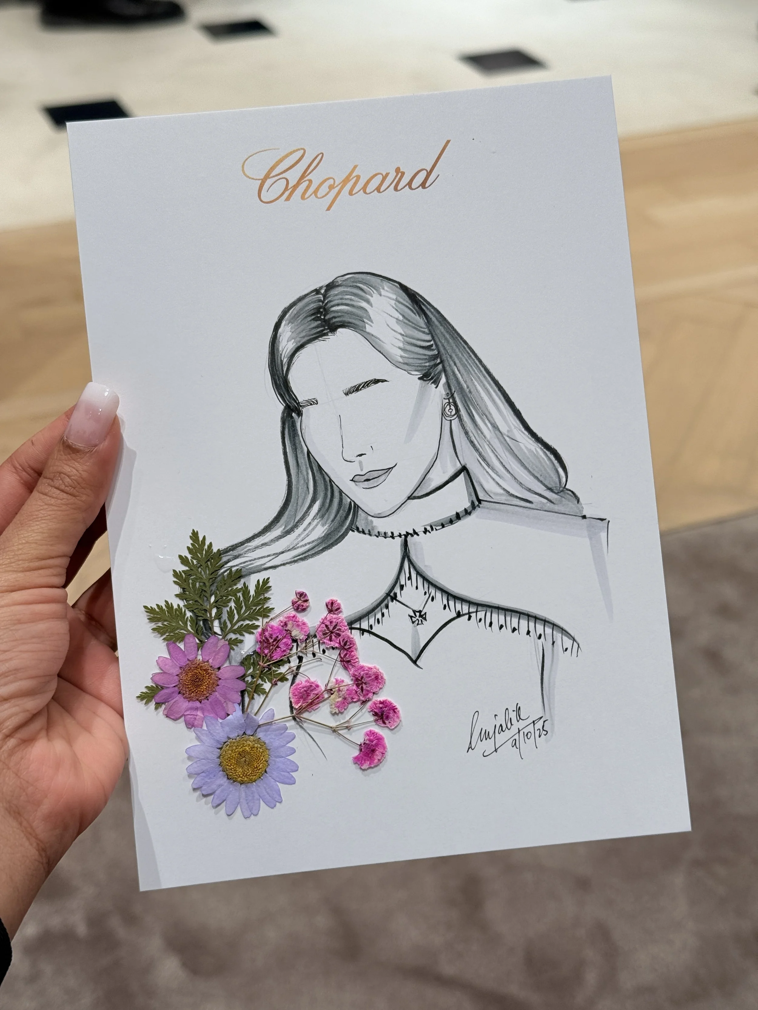 A hand holding a white card with a hand-drawn portrait of a woman with long hair and earrings, decorated with real pink, purple, and yellow flowers, and a signature with a date.
