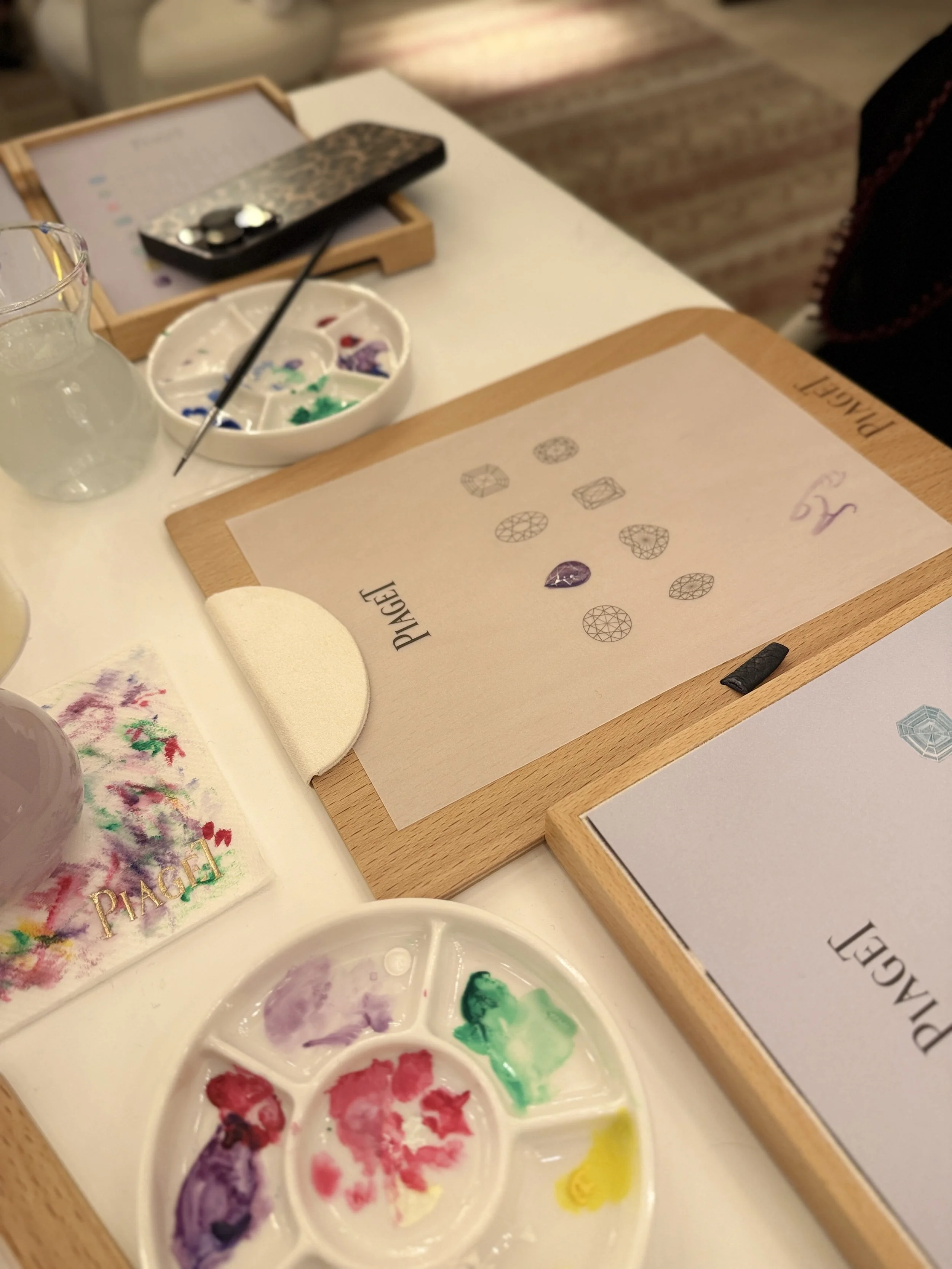 A watercolor palette with various colors, a glass pitcher, and a piece of paper with watercolor designs on a table.