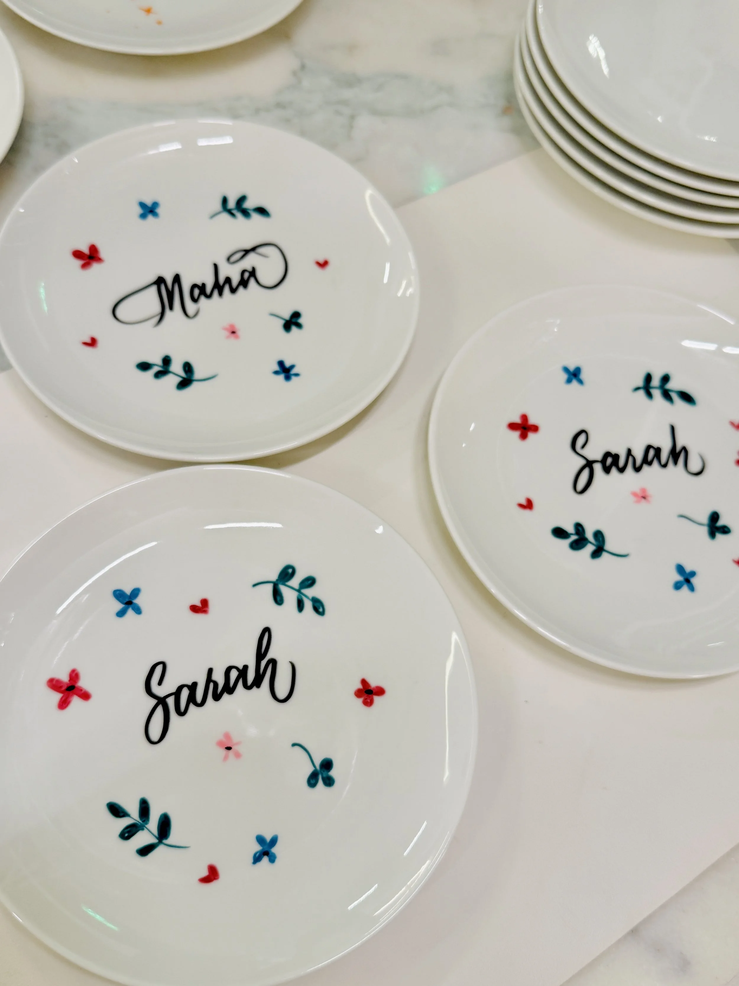 White party plates with colorful floral and heart designs, featuring personalized names 'Maha' and 'Sarah' written in black cursive.