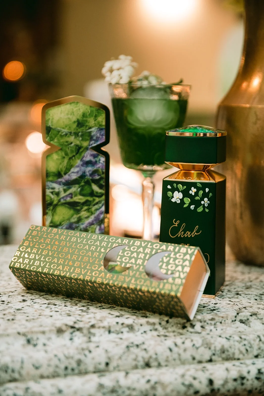 A green and gold Bulgari perfume box, a green bottle of perfume with floral design, and a decorative glass container with flowers and a green drink are displayed on a granite countertop.