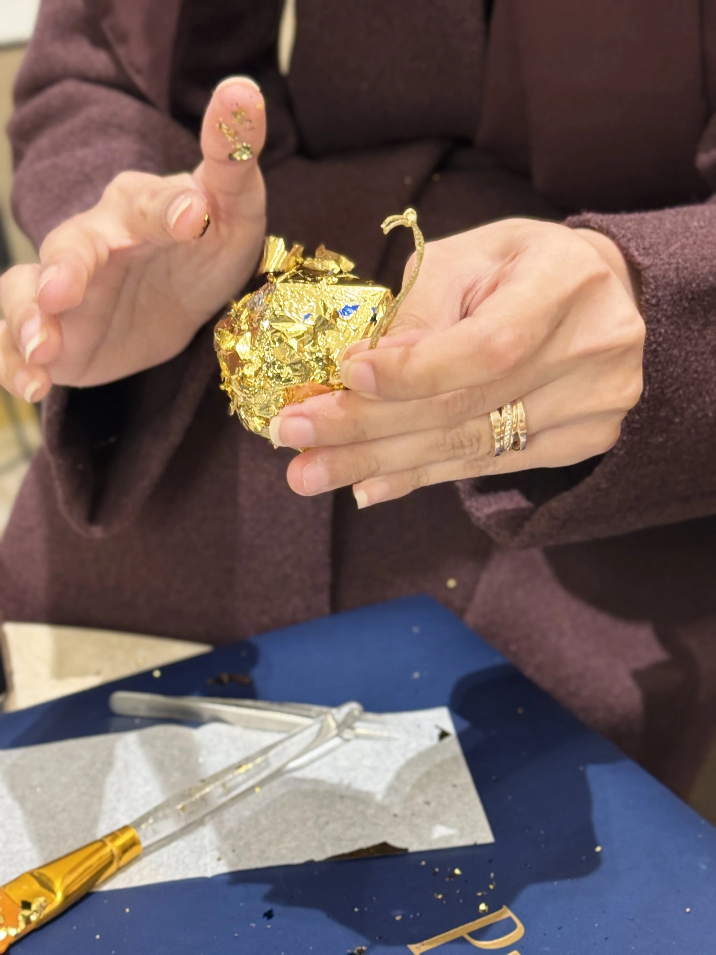 Person holding a crumpled gold-wrapped chocolate or gift, with finger tips decorated with gold glitter nail polish. On the table are tools, a paper, and scattered crumbs.