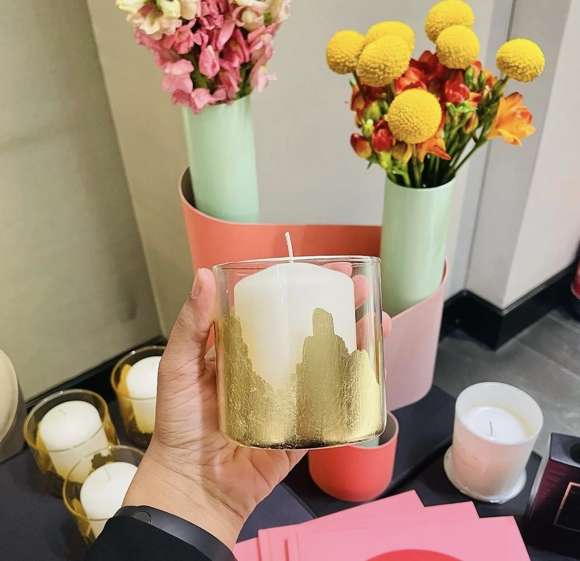 Person holding a white and gold candle in front of pink and green flower vases with pink and yellow flowers on a black surface.