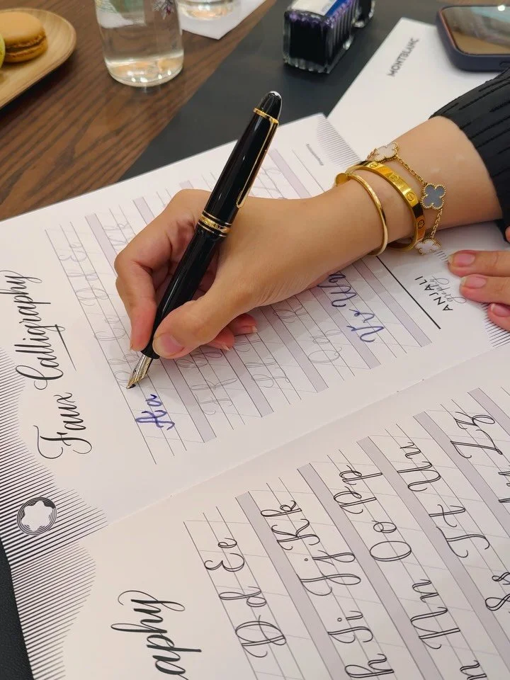 Person practicing calligraphy with a black pen on practice sheets, wearing gold bracelets, with a bottle of water, macaron, and stationery on a wooden table.