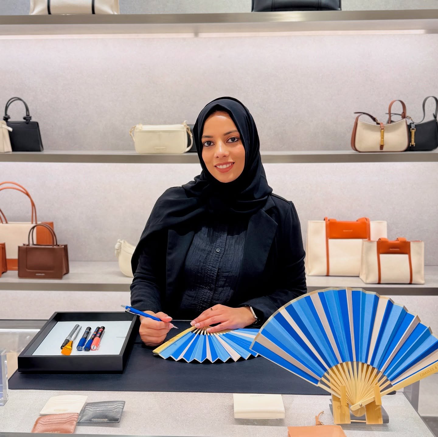 Woman with a black headscarf sitting at a table in a handbag store, holding a pen and a fan, with handbag displays in the background.