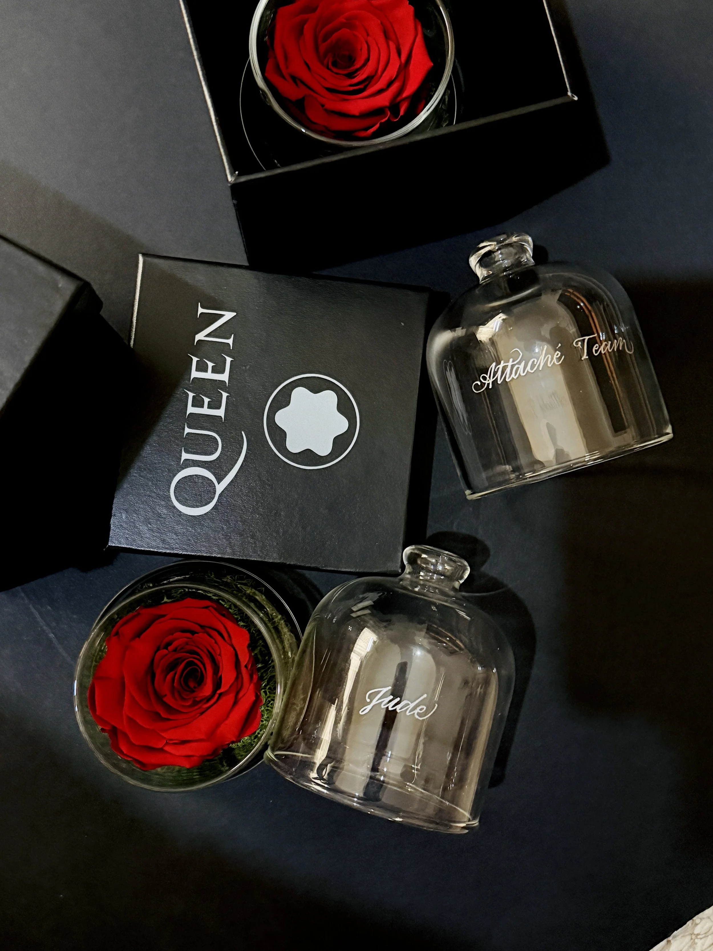 A black box with a red rose inside, and a clear bottle labeled "Attache Team" next to it on a dark surface.