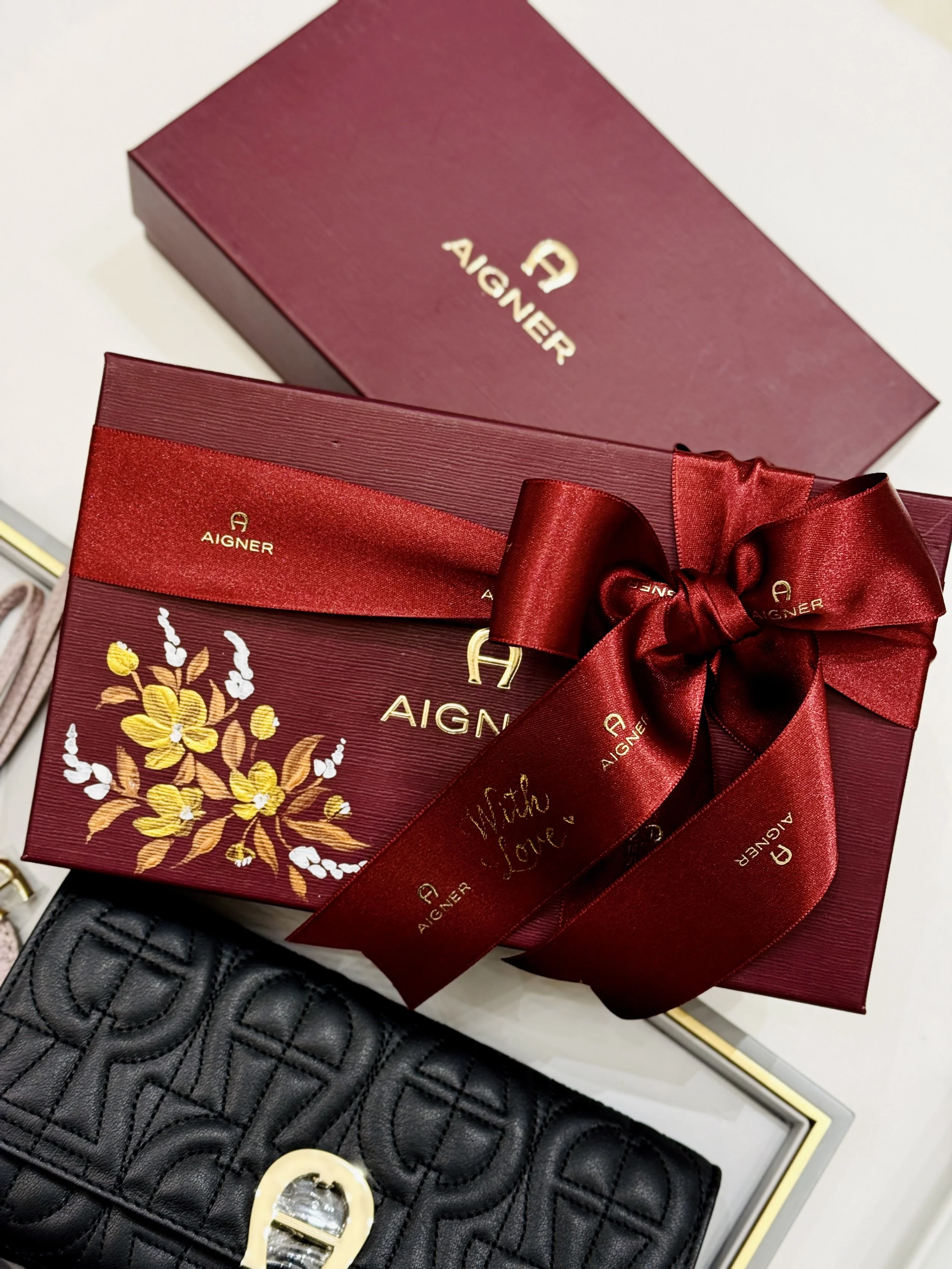 A maroon gift box with a floral design, a red satin ribbon with repeated 'Aigner' branding, a gray leather wallet with a gold 'A' emblem, and a jewelry box.