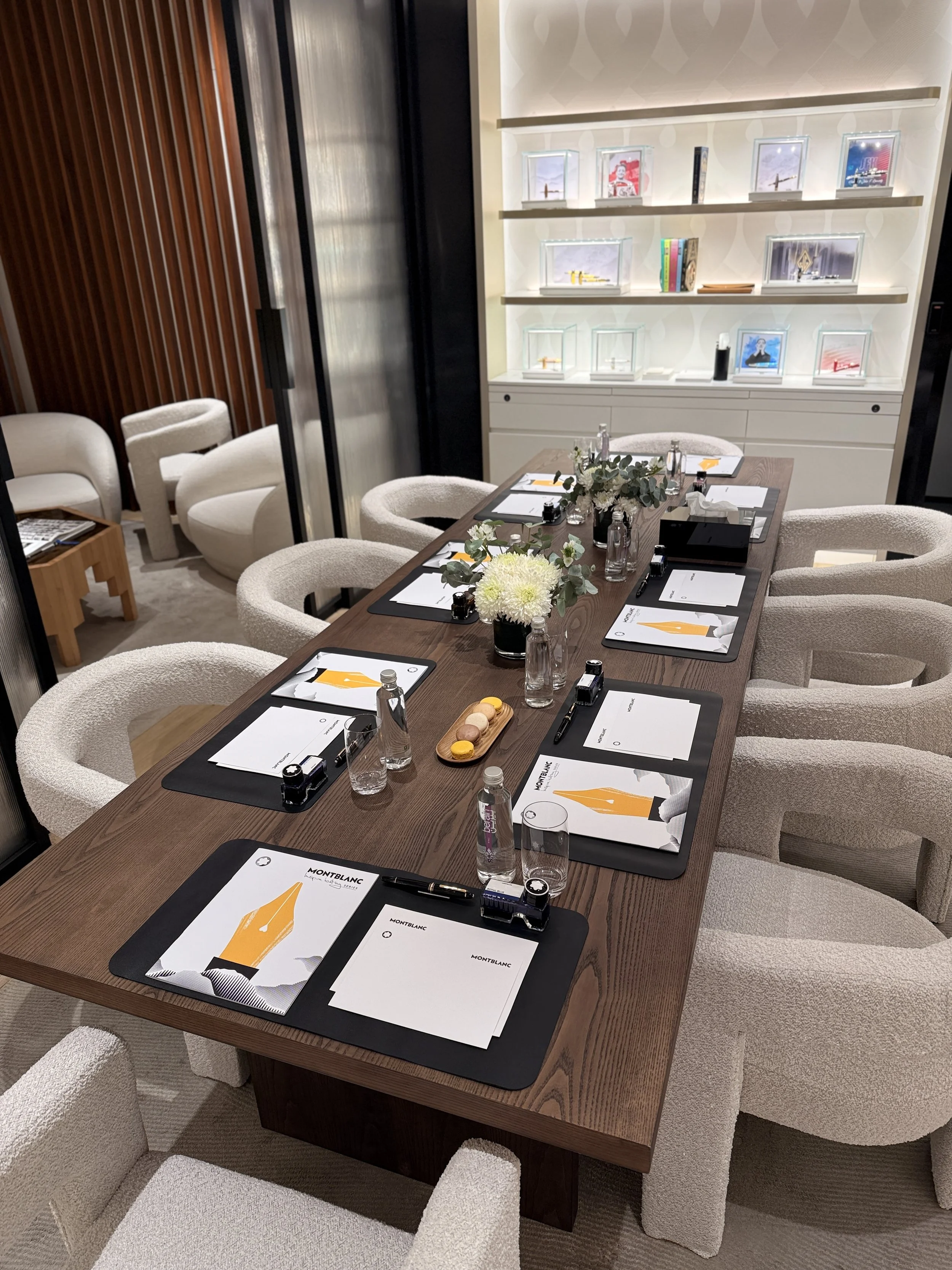 A conference room with a wooden table set for a meeting, surrounded by six white upholstered chairs. The table has notebooks, pens, water bottles, and a flower arrangement. In the background, there are built-in shelves with framed photographs, books,