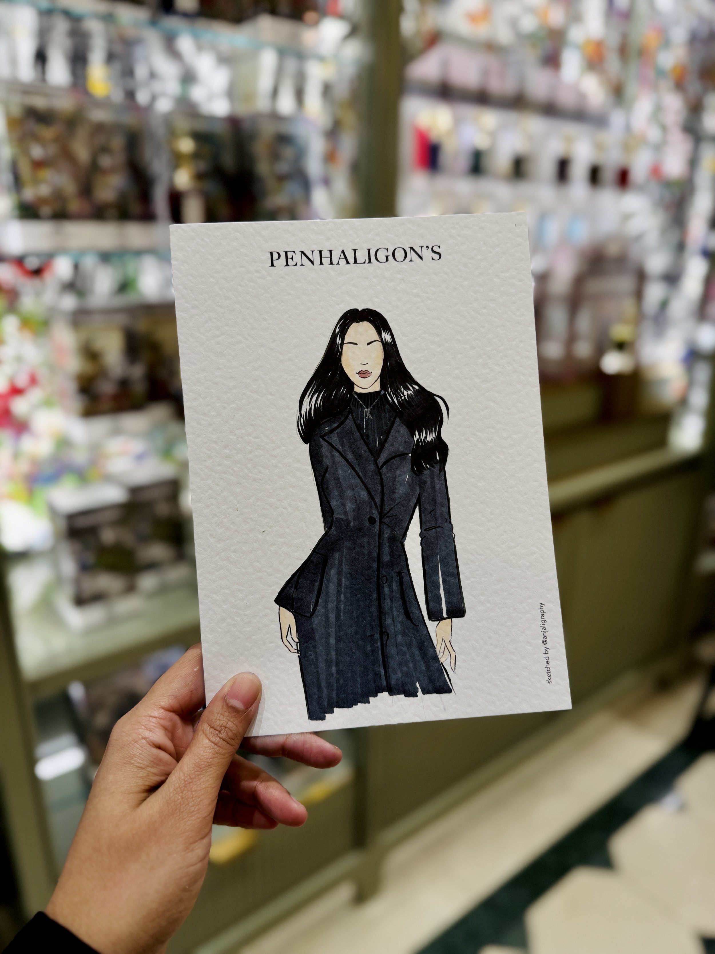 Illustration of a woman with long black hair wearing a black coat, on a textured white background with the heading 'PENHALIGON'S'. The background displays a grocery store aisle with various bottles and packages.
