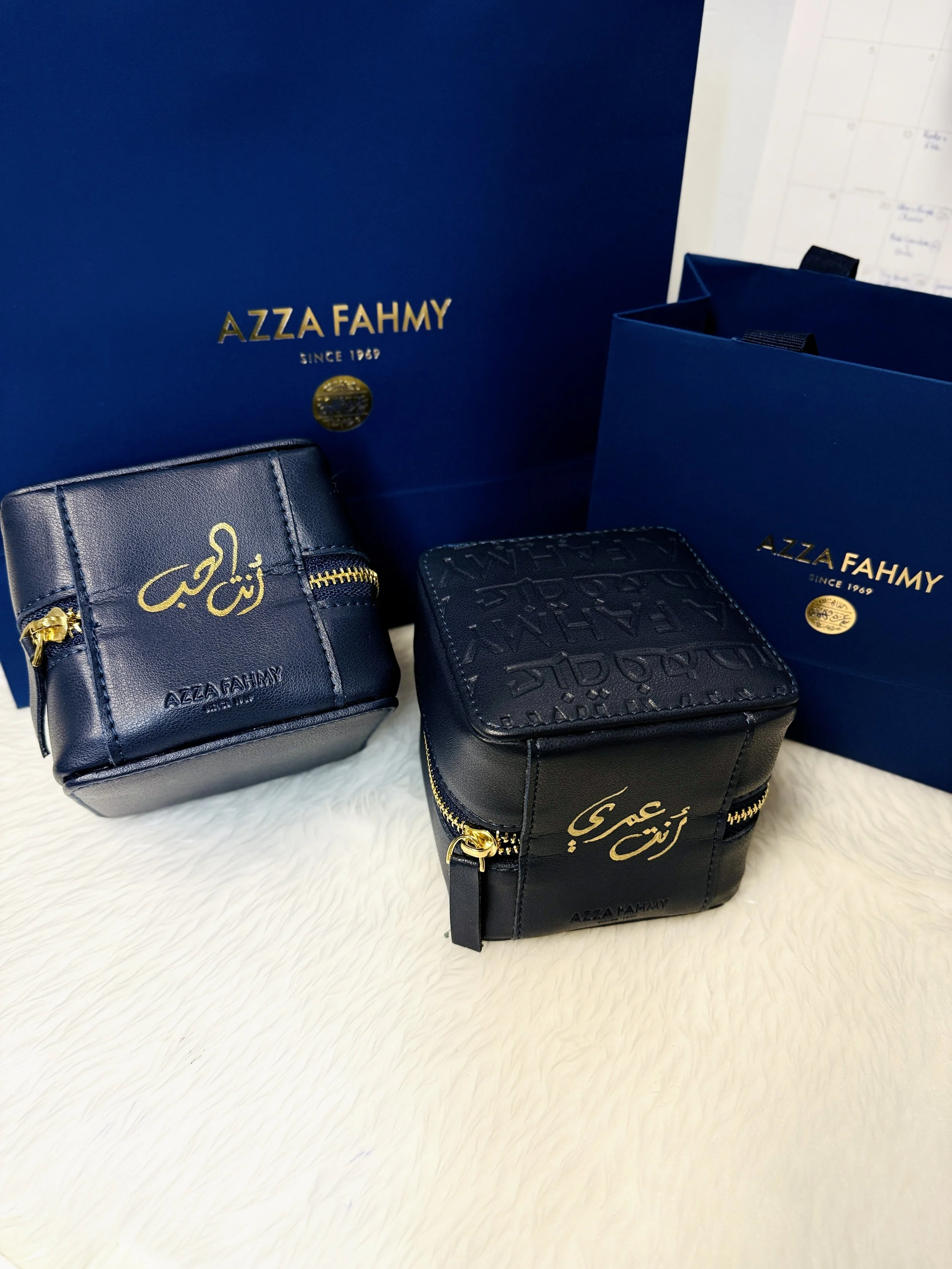 Three navy blue wallets with gold zipper accents and gold Arabic script, placed on a white furry surface. Two navy blue shopping bags with gold branding and text behind them.