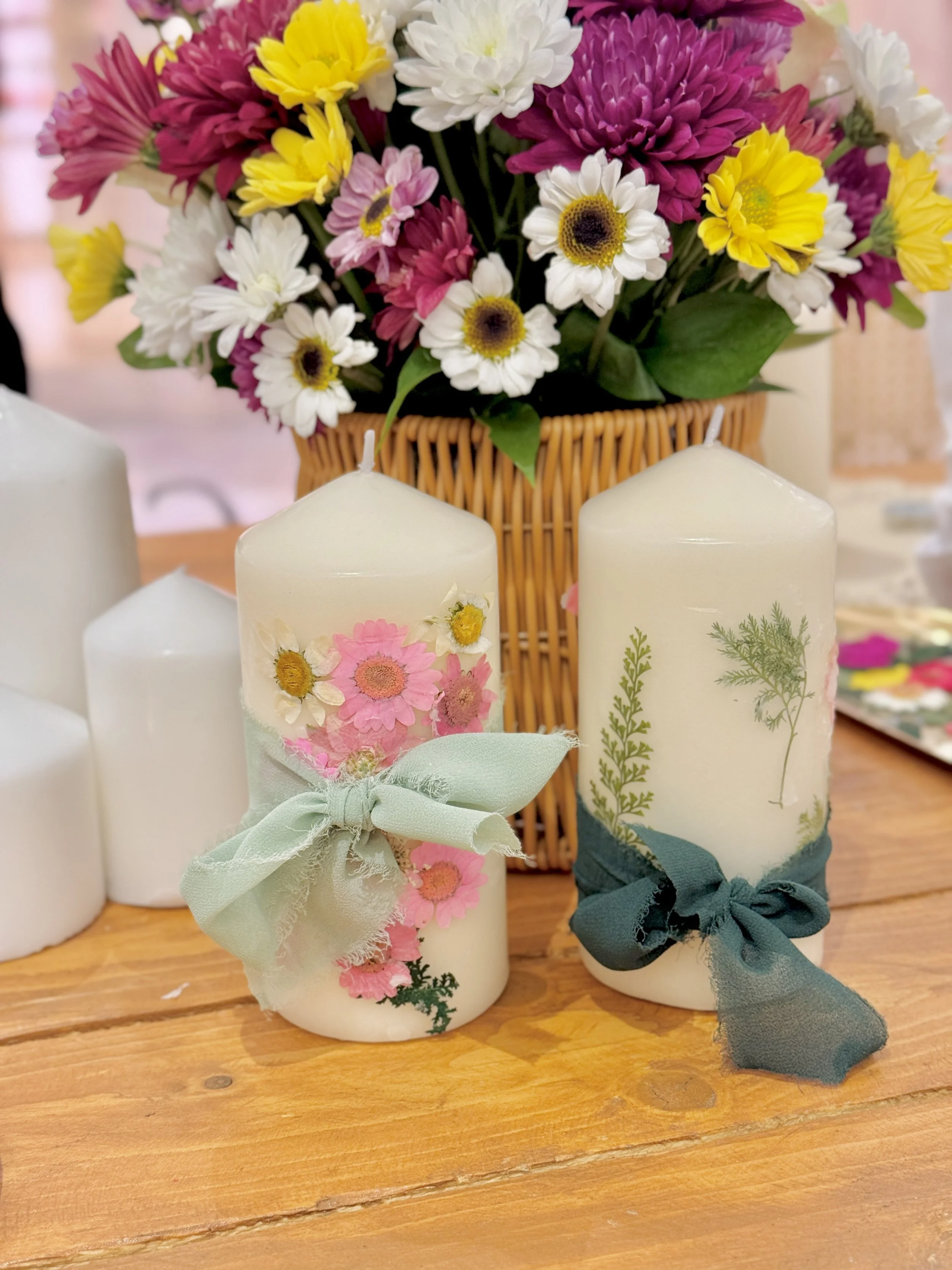 Three white candles decorated with pink, yellow, and green flower designs and tied with pastel-colored ribbons, placed on a wooden surface with a bouquet of colorful flowers in a wicker basket in the background.