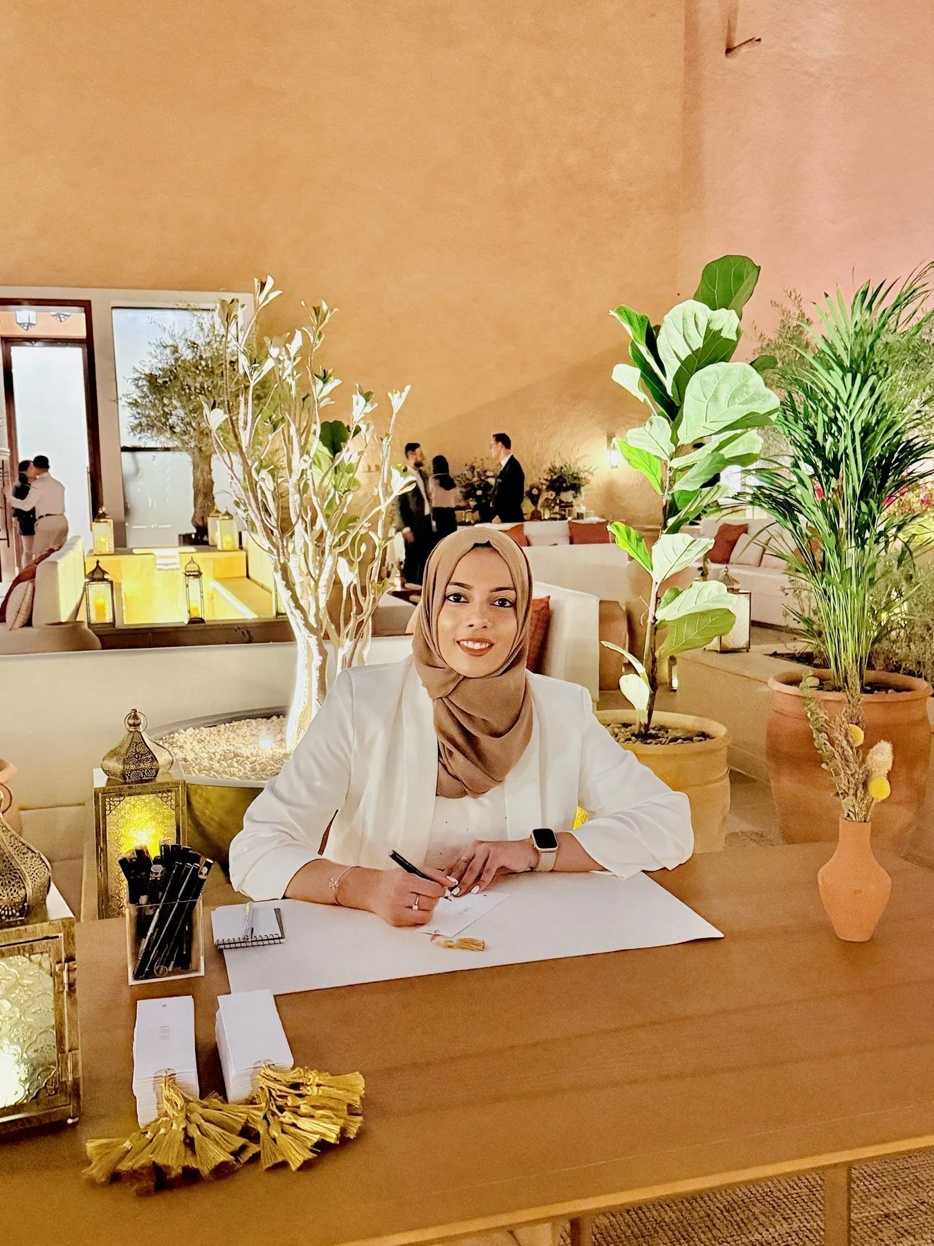 A woman wearing a hijab and white blazer sitting at a table in a warmly lit, decorated room with plants, lanterns, and a gold decorative item, holding a pen and smiling.