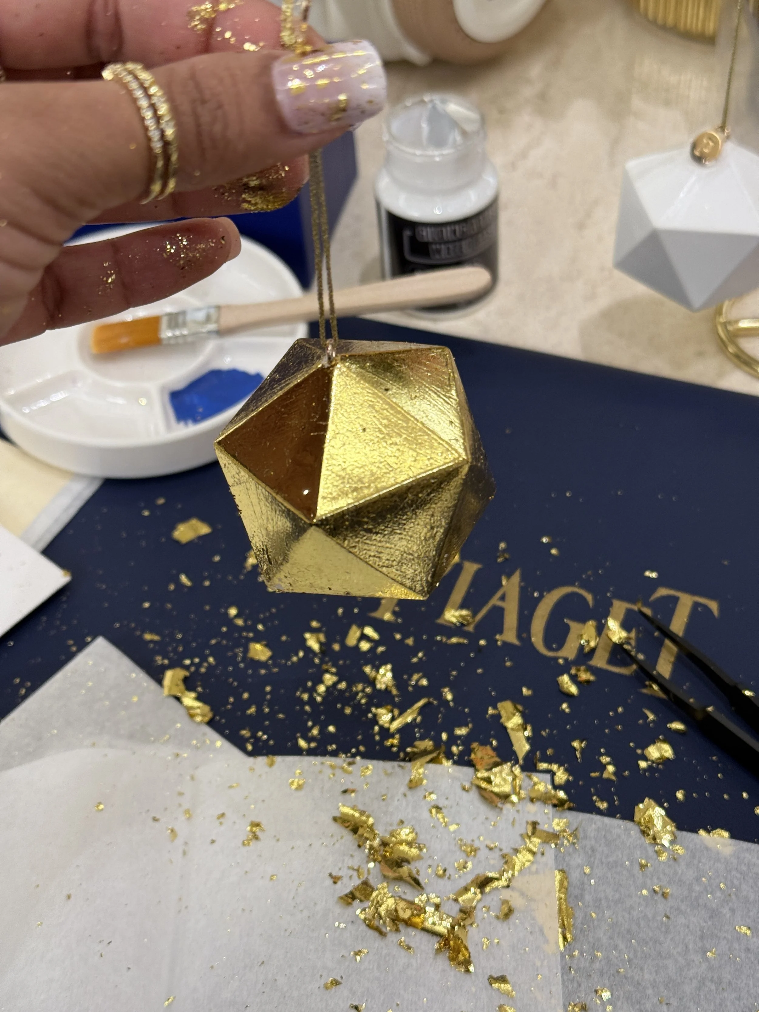 Hand holding a gold metallic geometric ornament with gold foil flakes scattered on the table and a background of various objects including a bottle of paint, a white container, and decorative items.