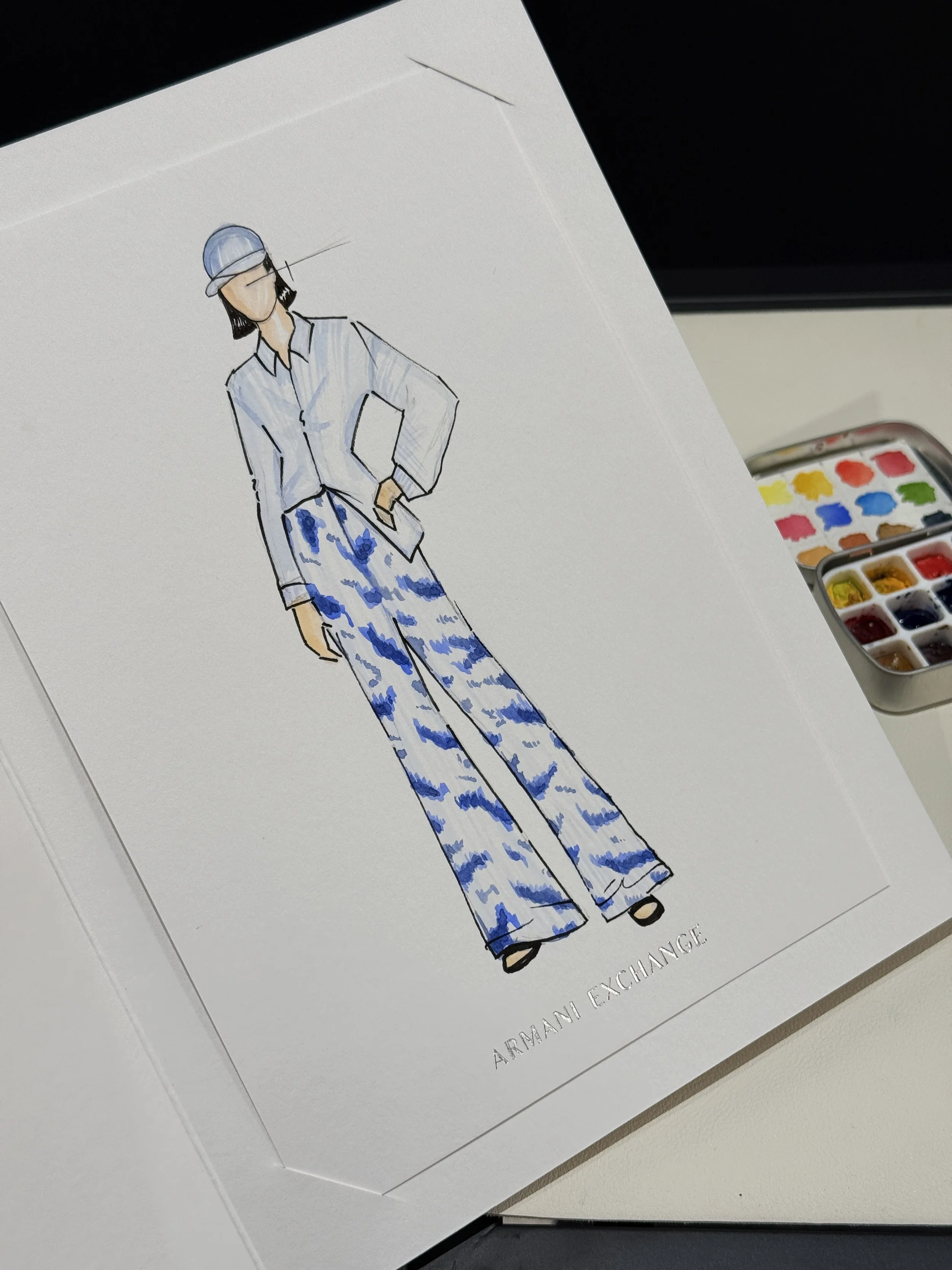 Fashion illustration of a woman wearing blue and white patterned wide-leg pants, a white blouse, a cap, and black shoes, with a watercolor palette visible beside the illustration.