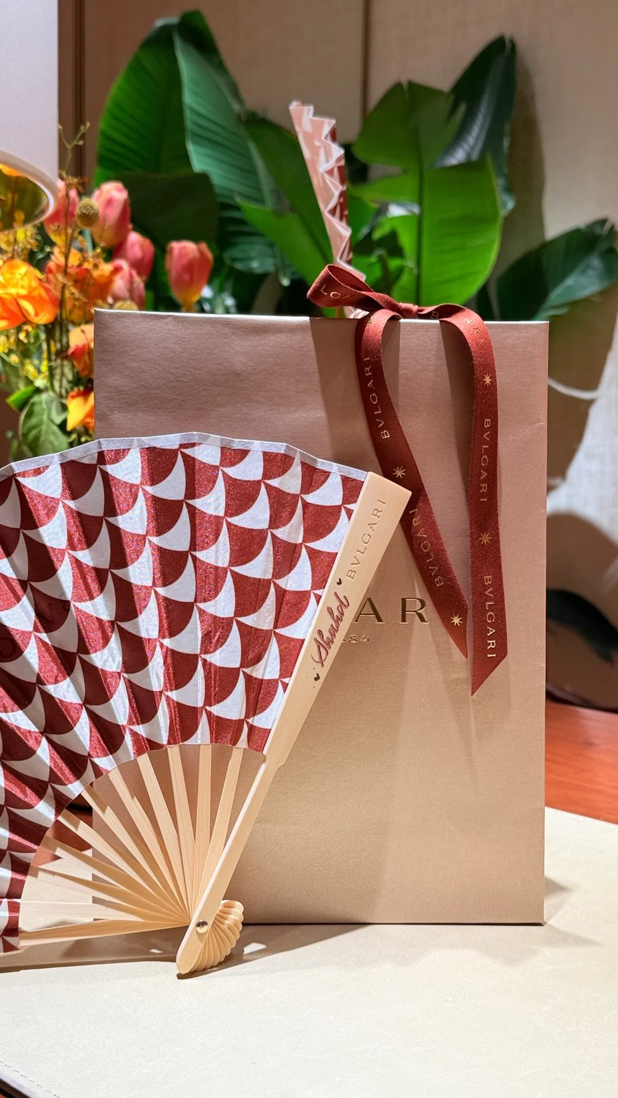 A pink Bvlgari shopping bag with a red ribbon, a small folding fan with a red and white pattern, and a bouquet of pink and orange flowers in the background.