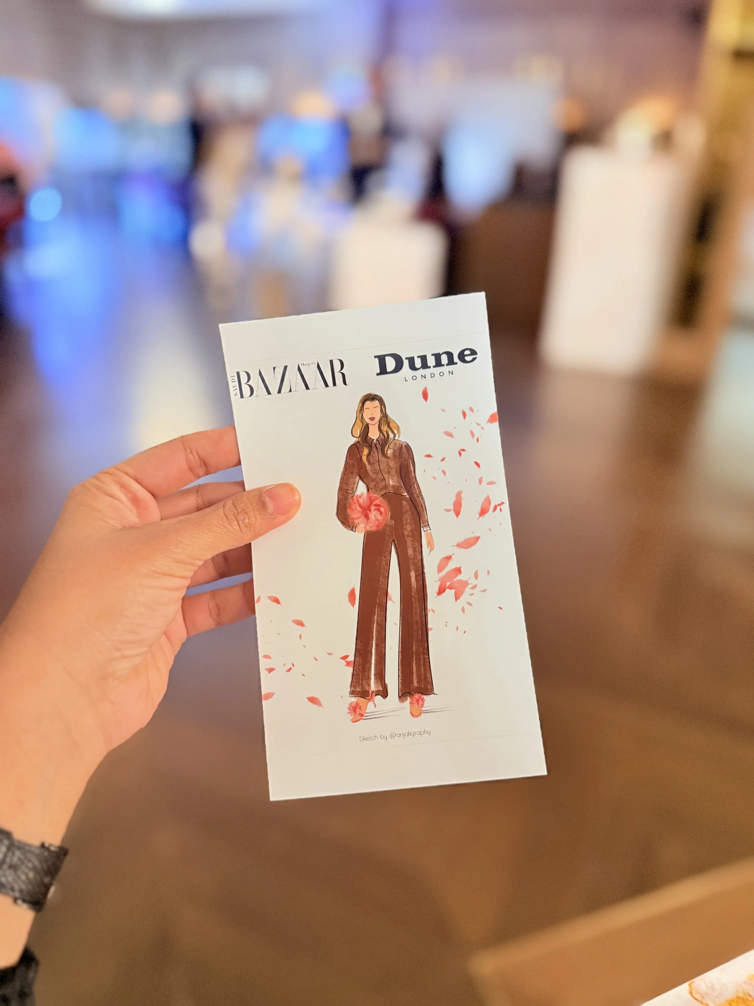 A hand holding a magazine page with an illustration of a woman in brown pants and brown jacket, holding a red flower, with falling red petals around her, and logos of Harper's Bazaar and Dune London at the top.
