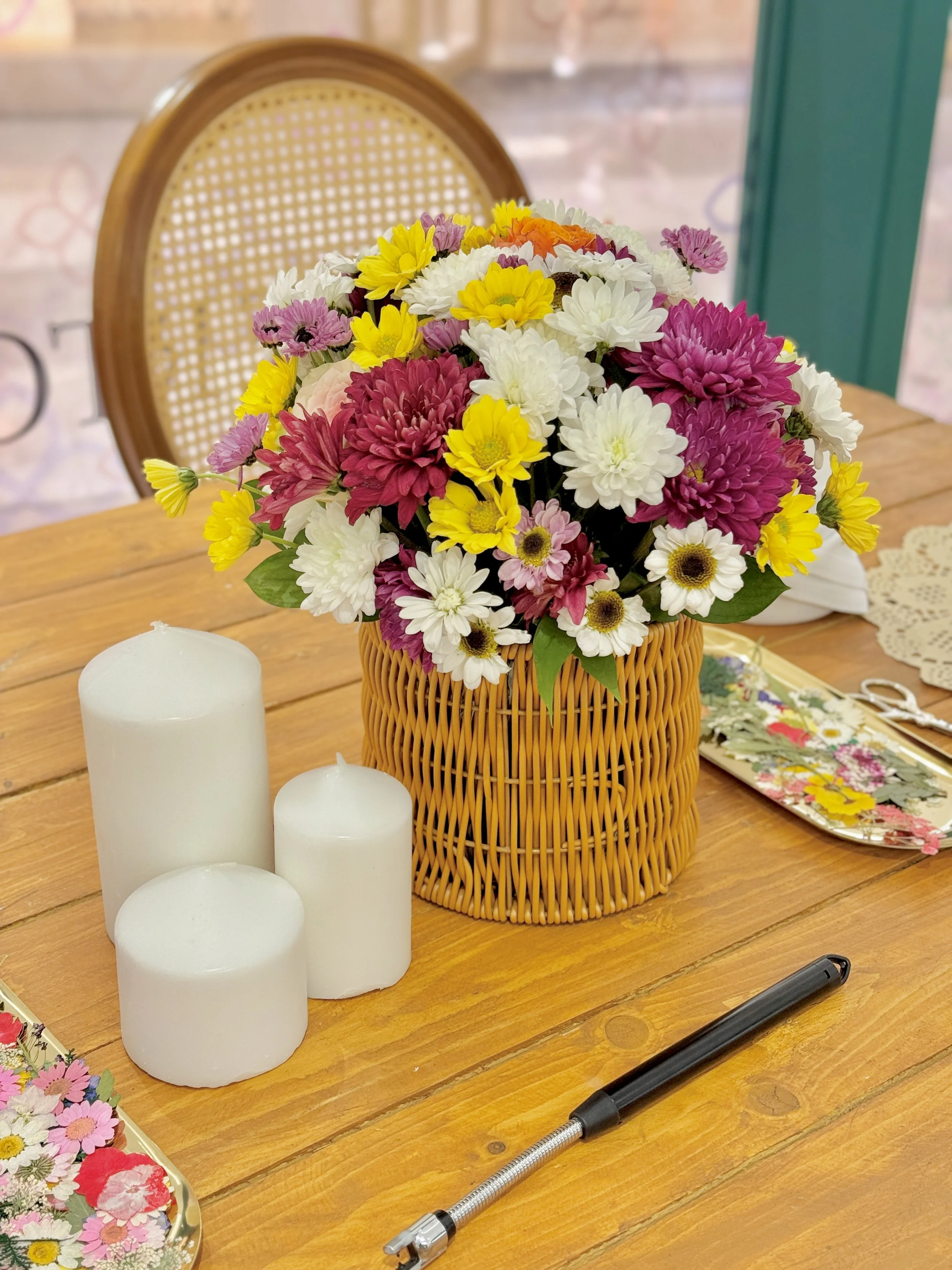A wicker basket filled with various colorful flowers on a wooden table, with three white candles, a pen, and some craft supplies nearby.
