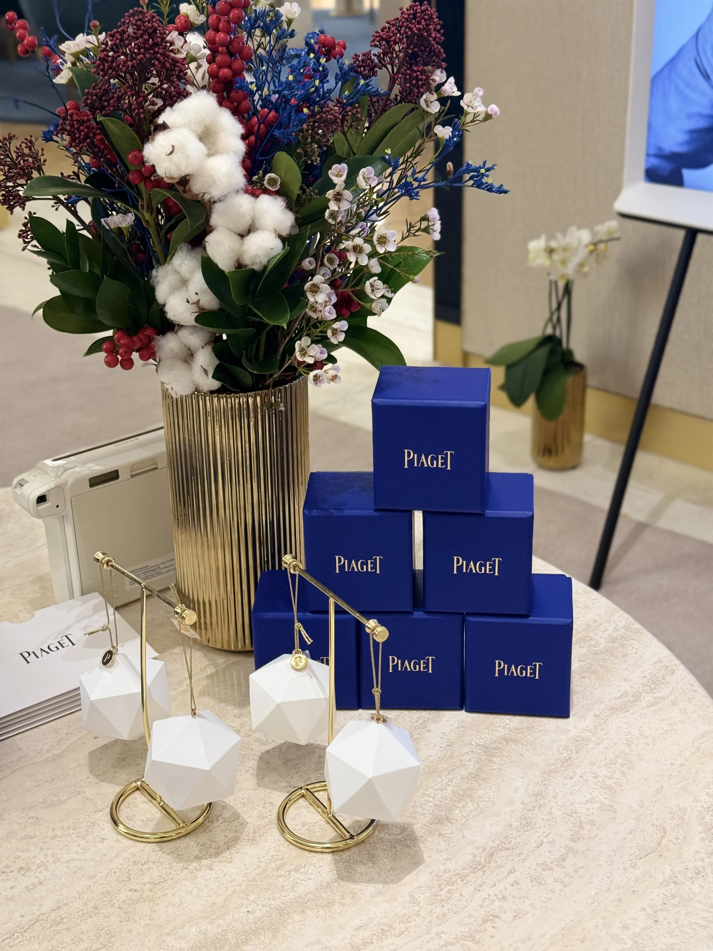 A jewelry display with yellow and white jewelry stands and a blue Piaget box. In the background, there is a vase with a bouquet of flowers including white, purple, red, and blue flowers with green leaves, placed in a gold-striped gold vase. There is 