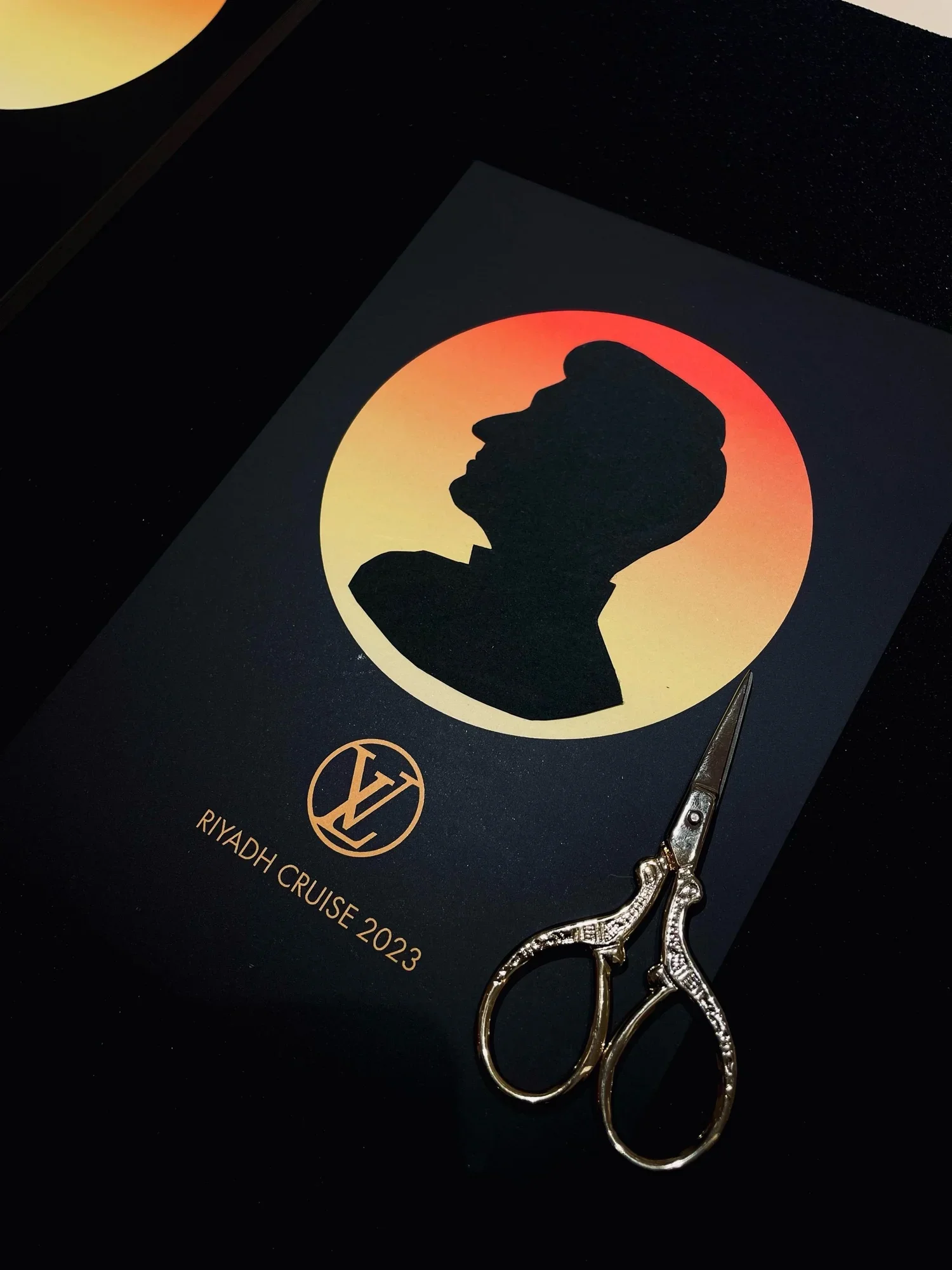 A black and gold card with a silhouette of a person's profile and the text 'RIYADH CRUISE 2023'. A pair of ornate scissors is placed on the card.