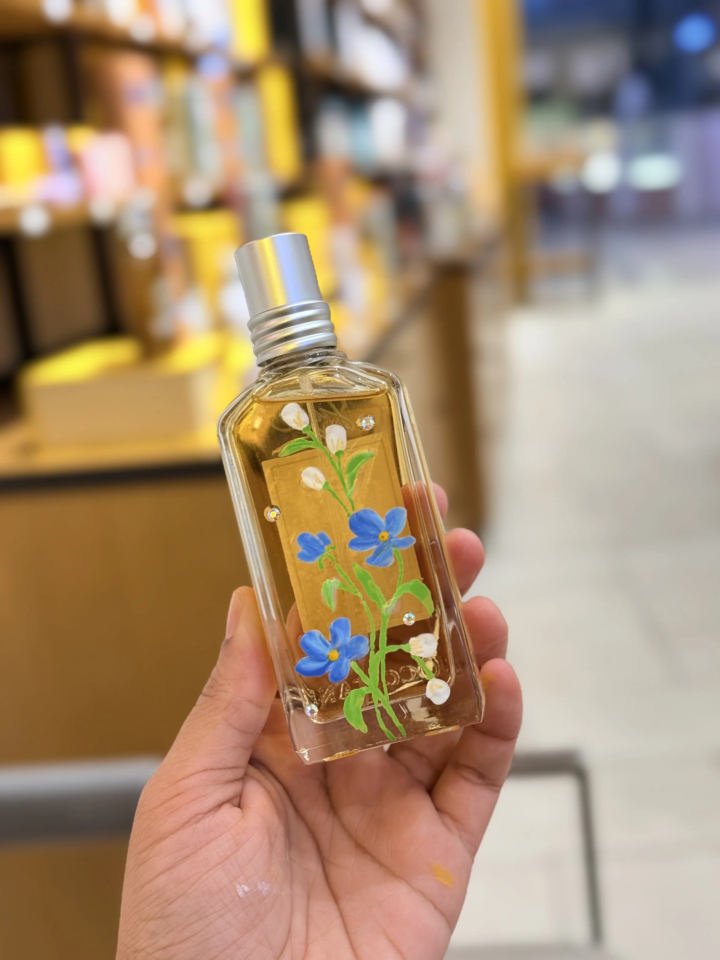 Hand holding a small spray bottle with blue flowers and white flowers on it, in a store with shelves in the background.