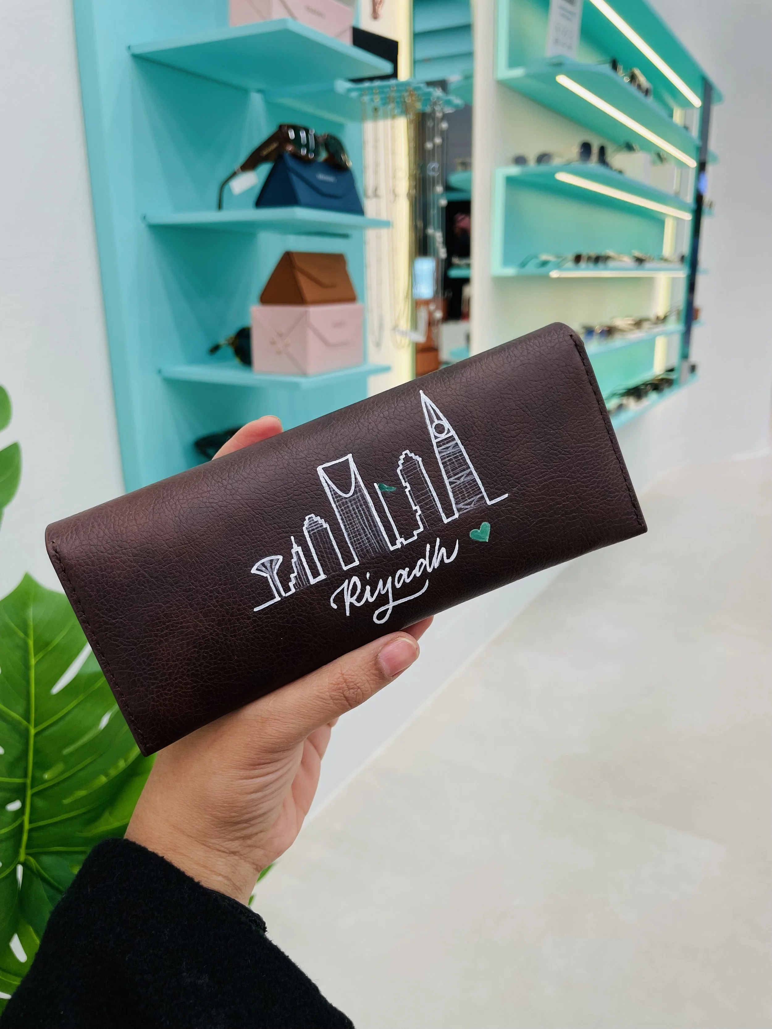 Hand holding a brown wallet with cityscape and text that says 'Riyadh' against a background of store shelves.