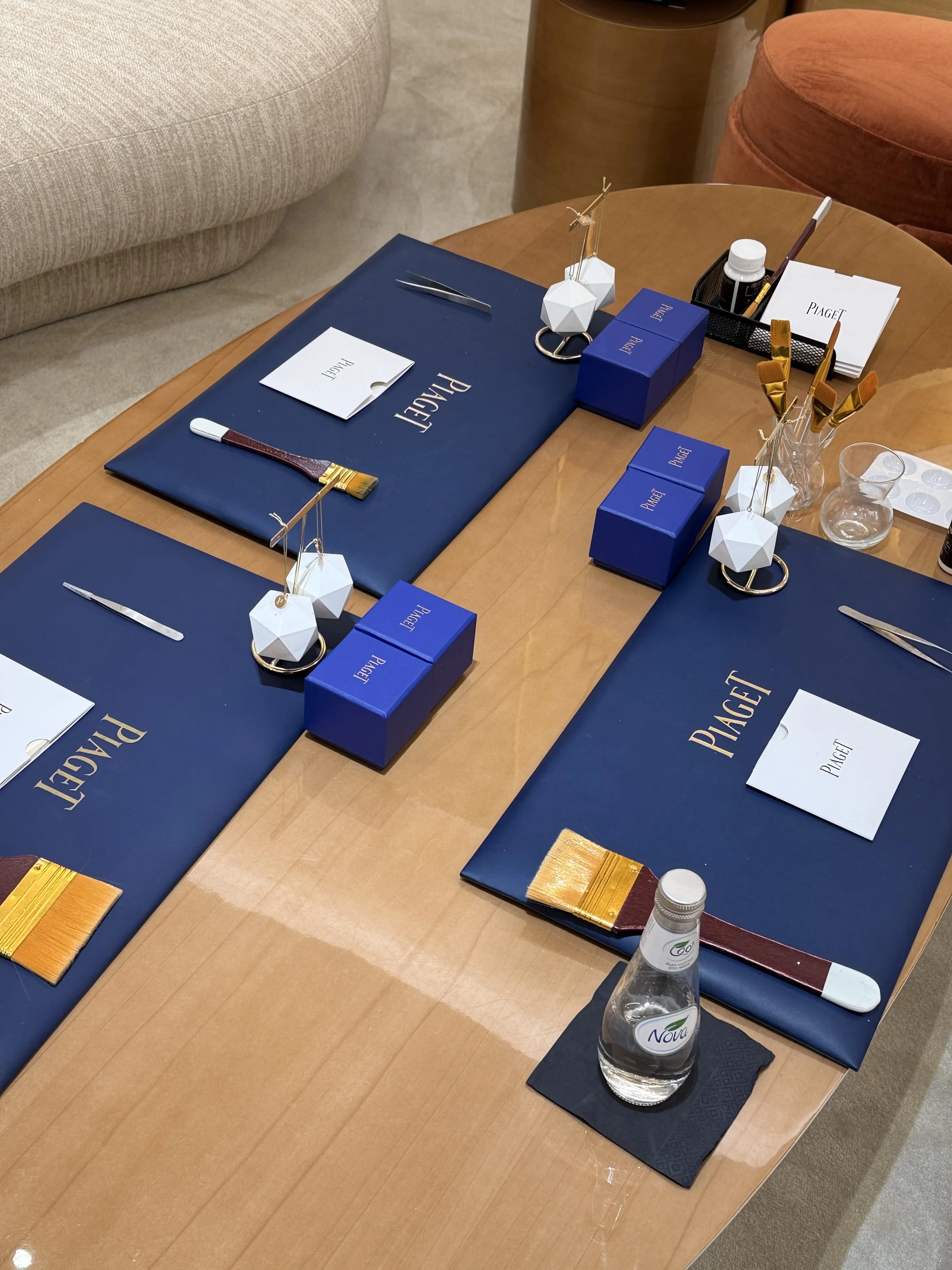 A wooden table set with blue placemats, white and blue Piaget presentation boxes, a small white booklet labeled 'Piaget,' small bottles of what appears to be medication, laboratory tools, and decorative items such as geometric vases with gold-colored