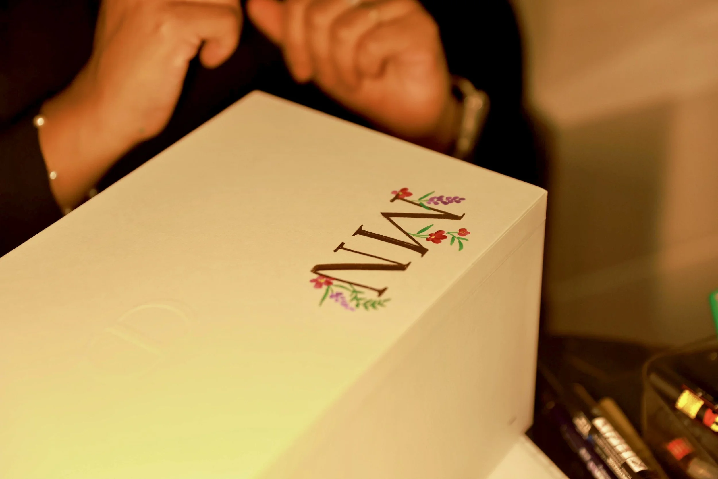 A white box with the word "WINE" written on it, decorated with small colorful flowers, and a person's hand holding a phone nearby.