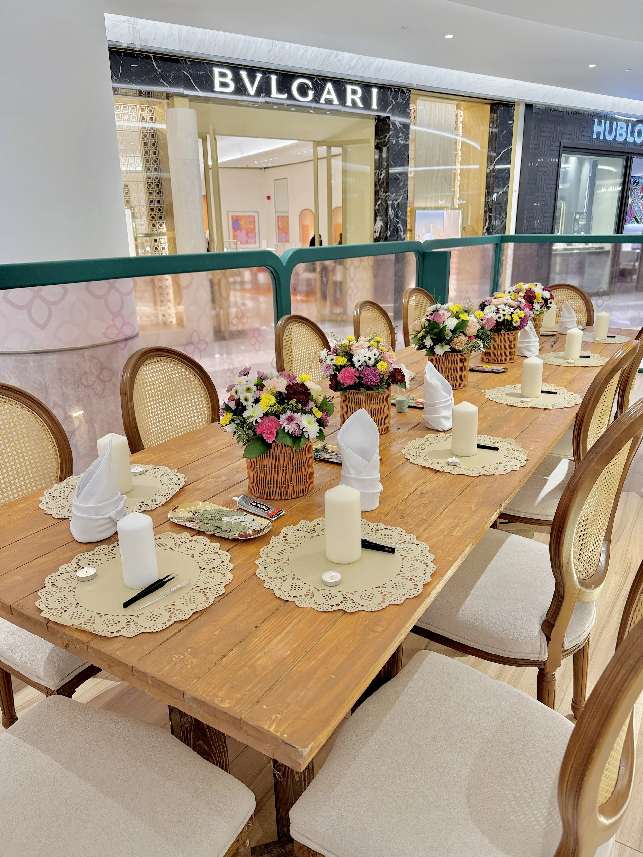 A dining table set with flower arrangements, candles, lace doilies, and napkins in a shopping mall near a Bvlgari store.