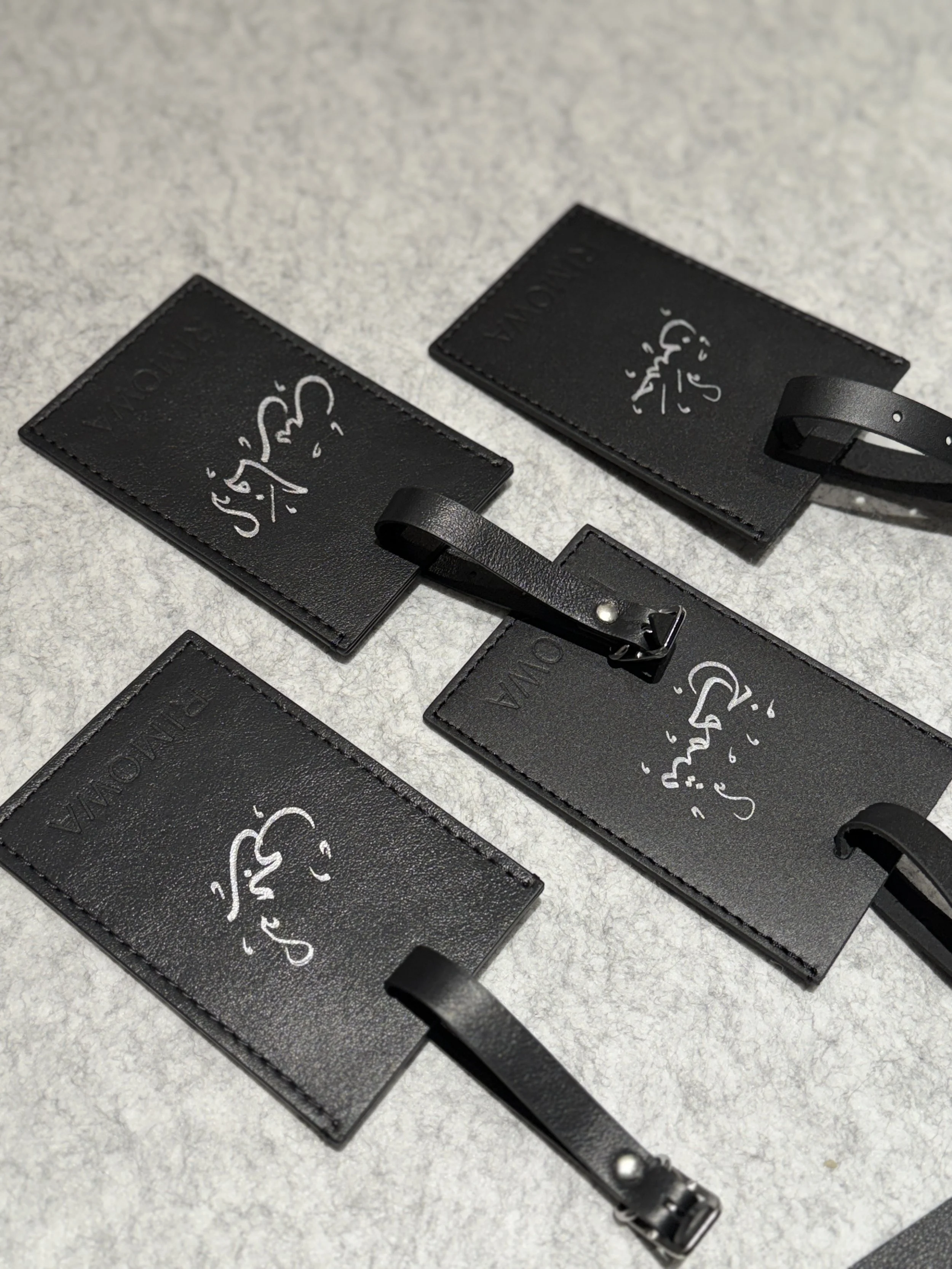 Close-up of black leather luggage tags with white handwritten-style text and graphics, attached to black leather straps, on a light gray textured surface.