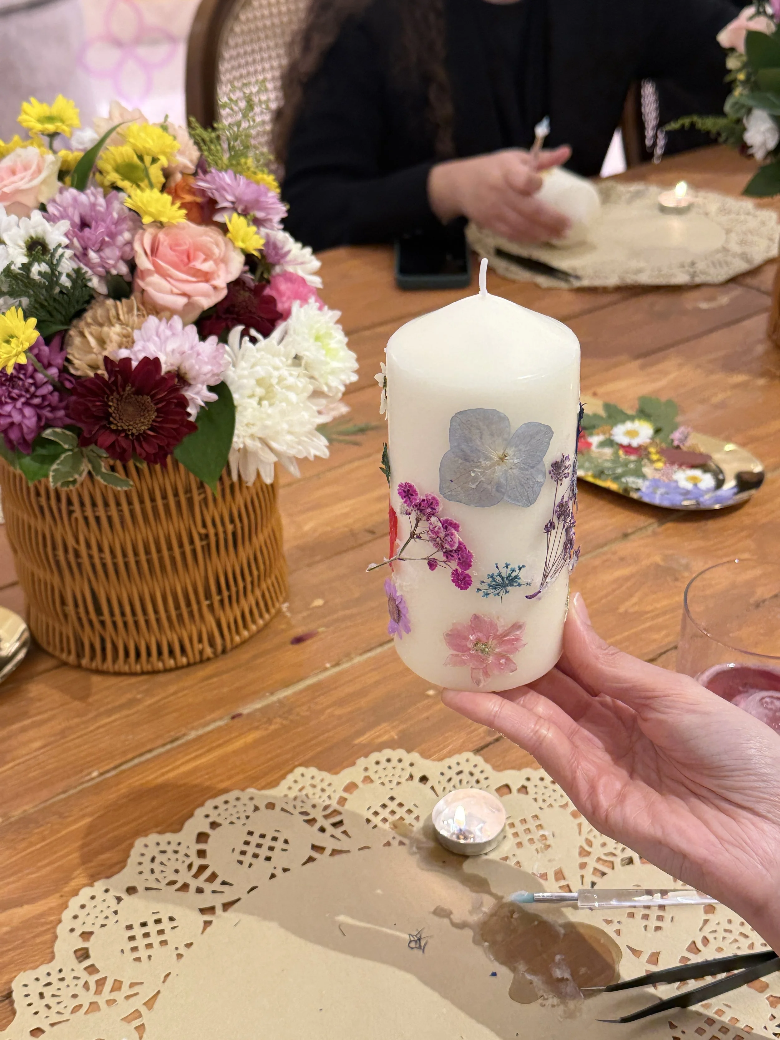 Person holding a decorated white candle with pressed flowers, table with flowers, candle, and lace doily in the background.