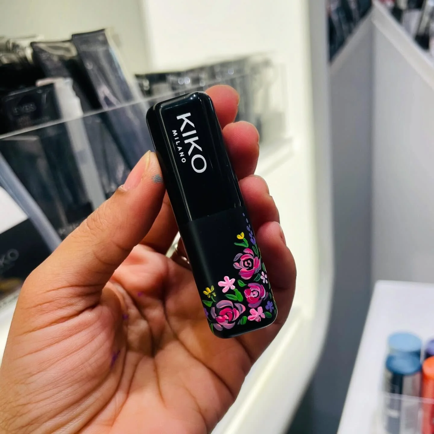 Hand holding a black Kiko Milano lip gloss with a floral design on the lower half of the packaging, showing the Kiko Milano logo on the upper part.