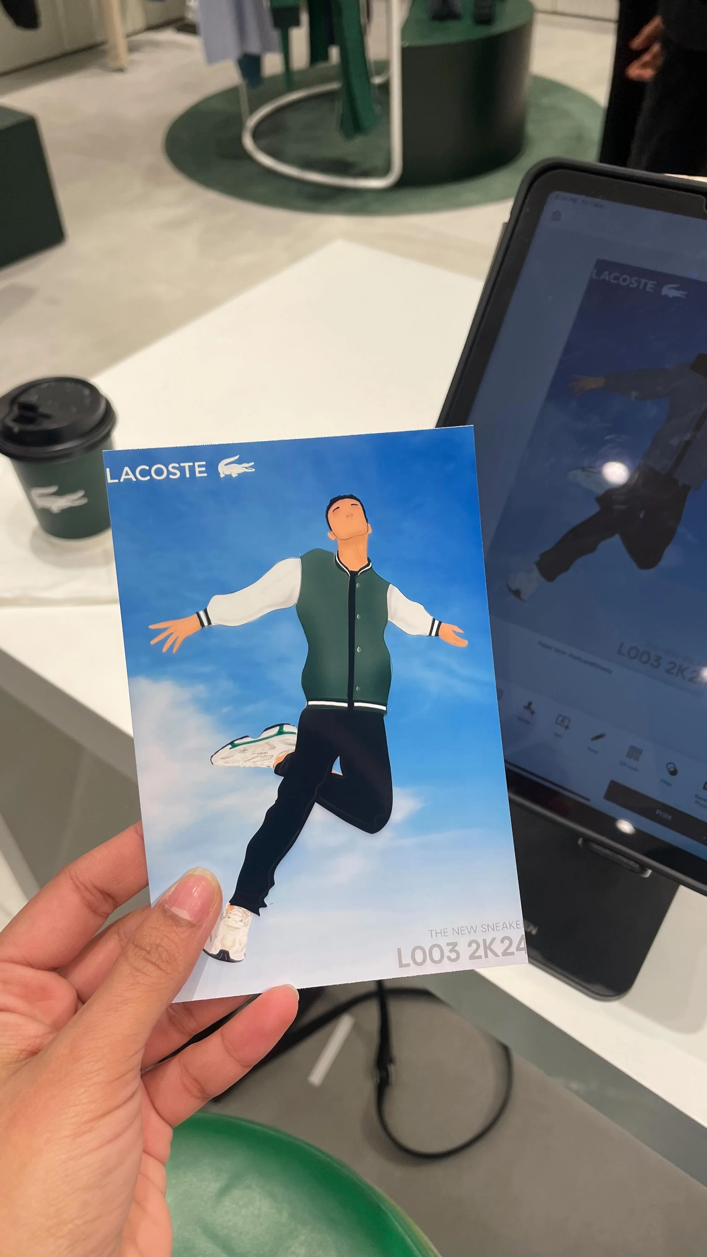 A person holding a Lacoste advertisement card with an animated illustration of a person in a green varsity jacket with outstretched arms, standing against a partly cloudy sky background. There is a coffee cup and a tablet on a table in the background