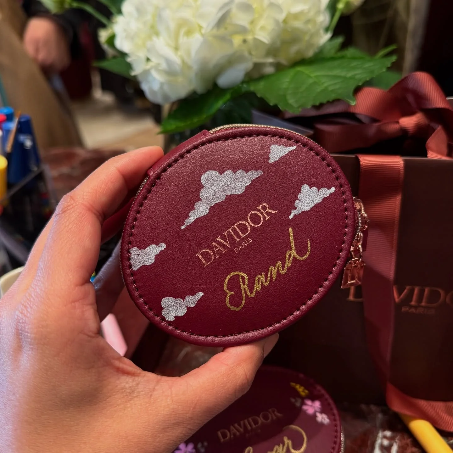A hand holding a red circular coin purse with white clouds, gold text reading 'DAVIADOR PARIS' and a handwritten gold name 'Ramau'. In the background, there are white flowers and a brown gift box with a pink bow.