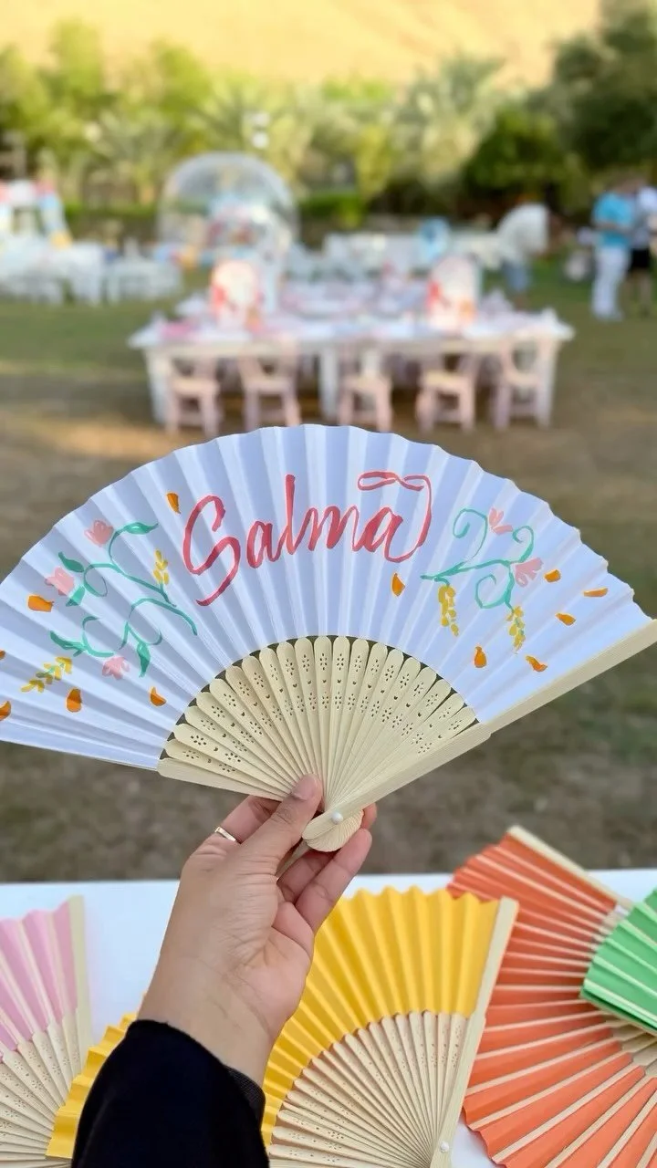 Hand holding a decorated festival fan with the word 'Salma' on it, with outdoor tables and chairs in the background, indicating an outdoor celebration or event.