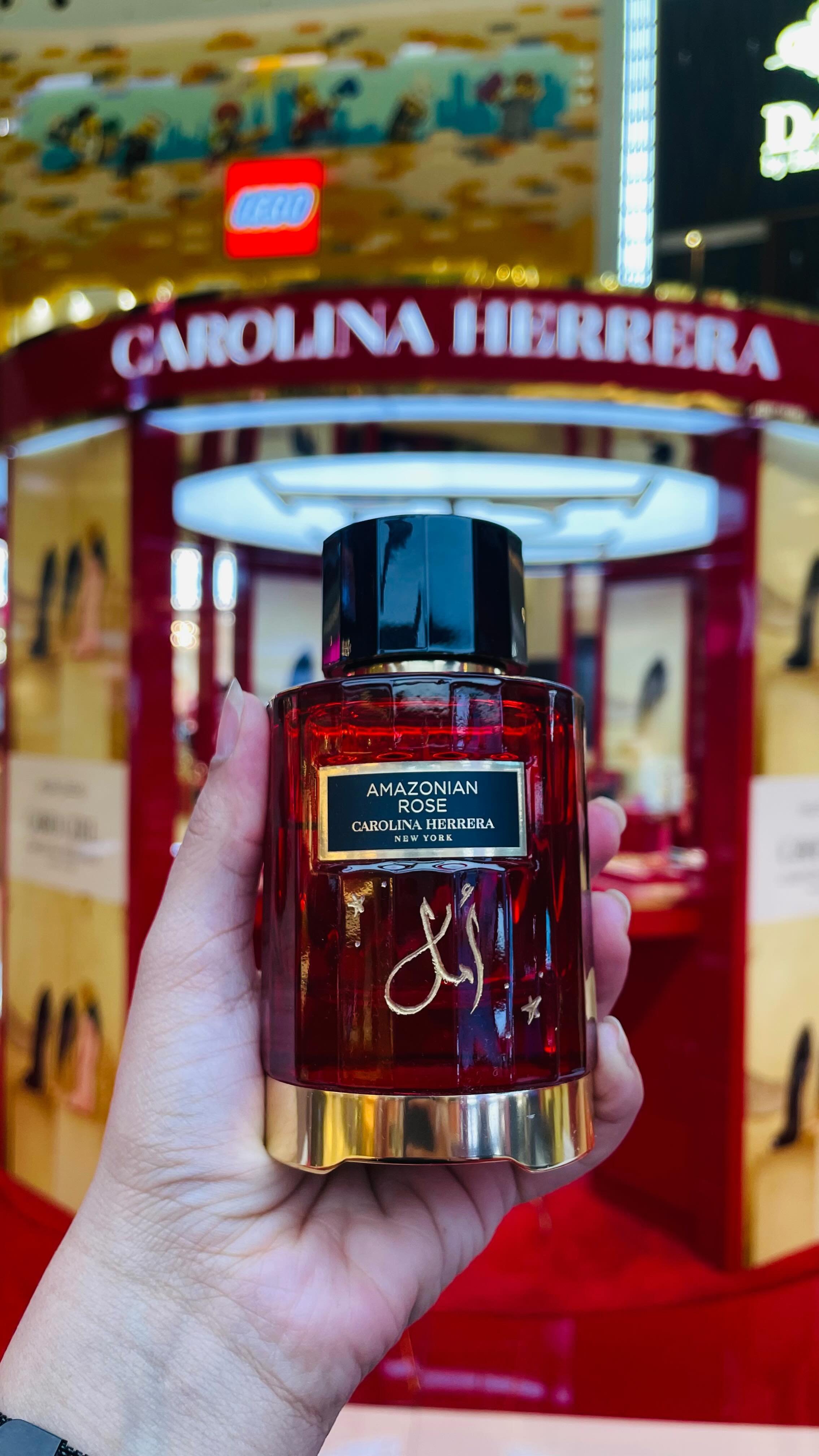 A hand holding a bottle of Carolina Herrera Amazonian Rose perfume in front of a red and gold stand with the name prominently displayed. The background includes a LEGO store sign and yellow displays.